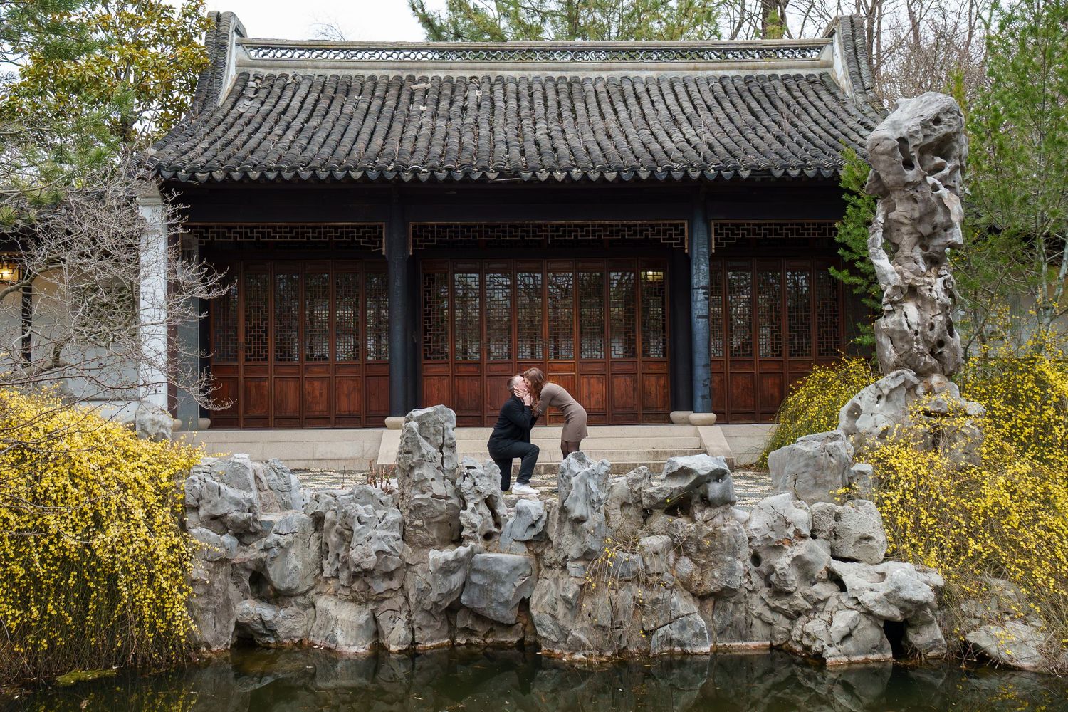 Couple embracing on decorative rocks by a pond in front of a Chinese pavilion.