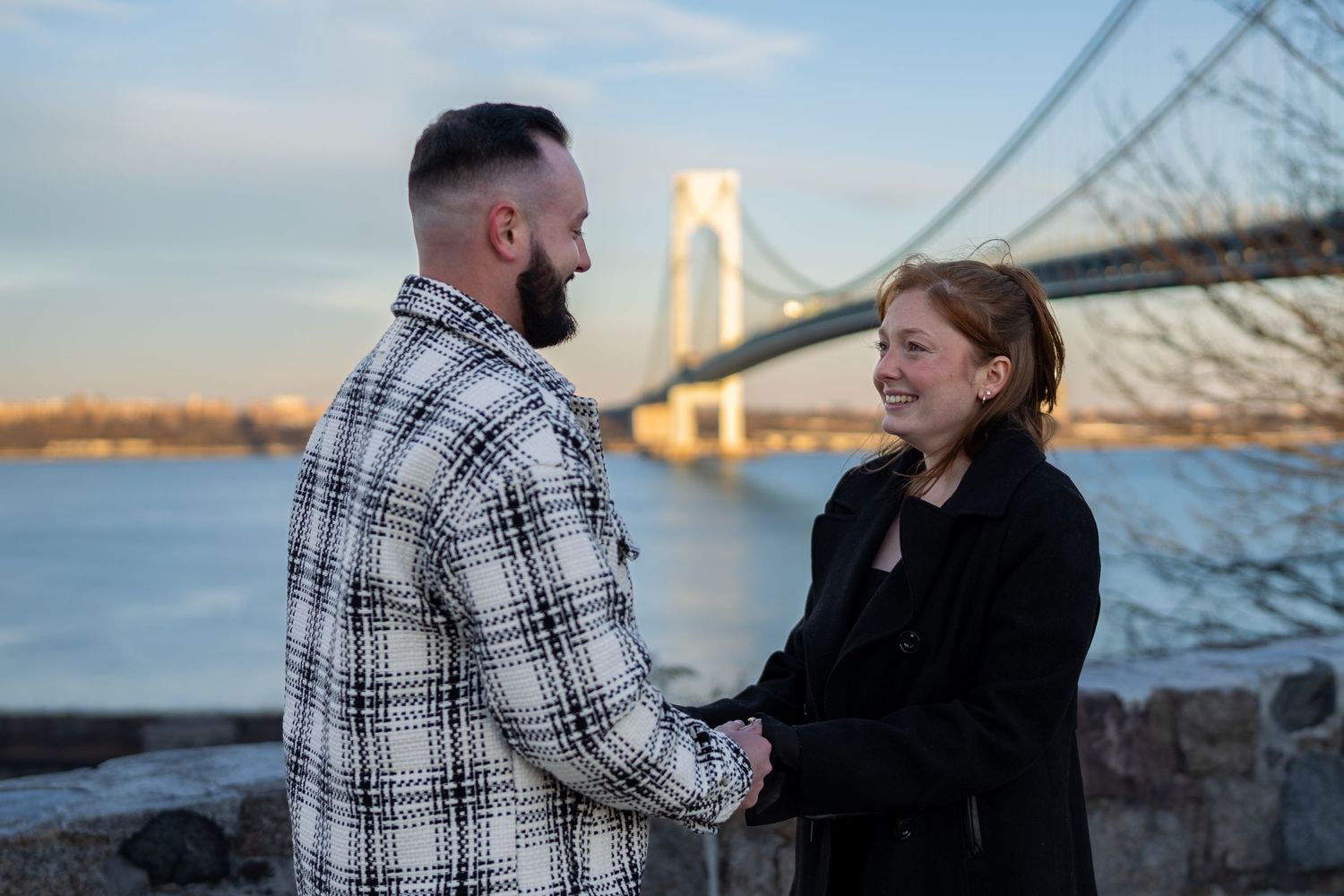 Smiling couple holding hands facing each other with the Verrazzano-Narrows Bridge at sunset.