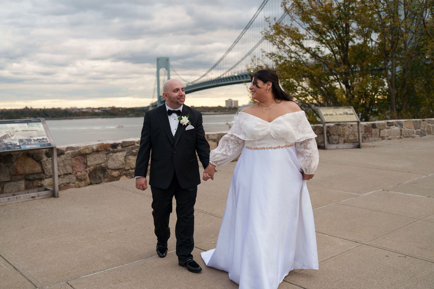 Wedding couple walks hand-in-hand on waterfront plaza with iconic suspension bridge backdrop.