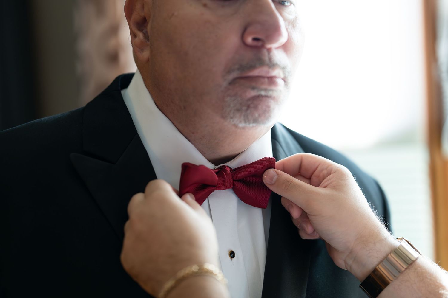 A man in a tuxedo has his red bow tie adjusted by another person's hands before a wedding.