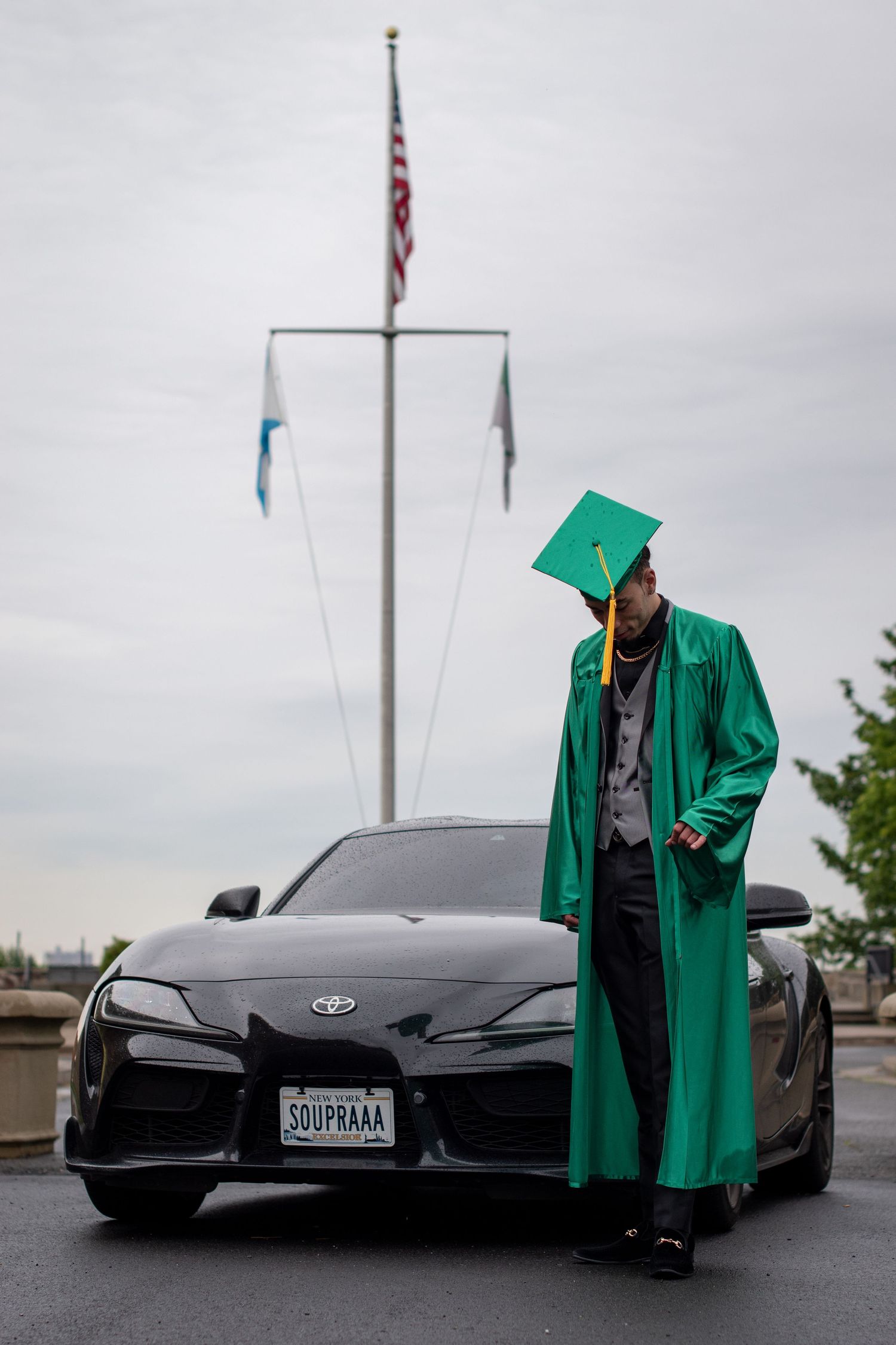 A graduate in green cap and gown poses beside a black Toyota Supra under cloudy skies.