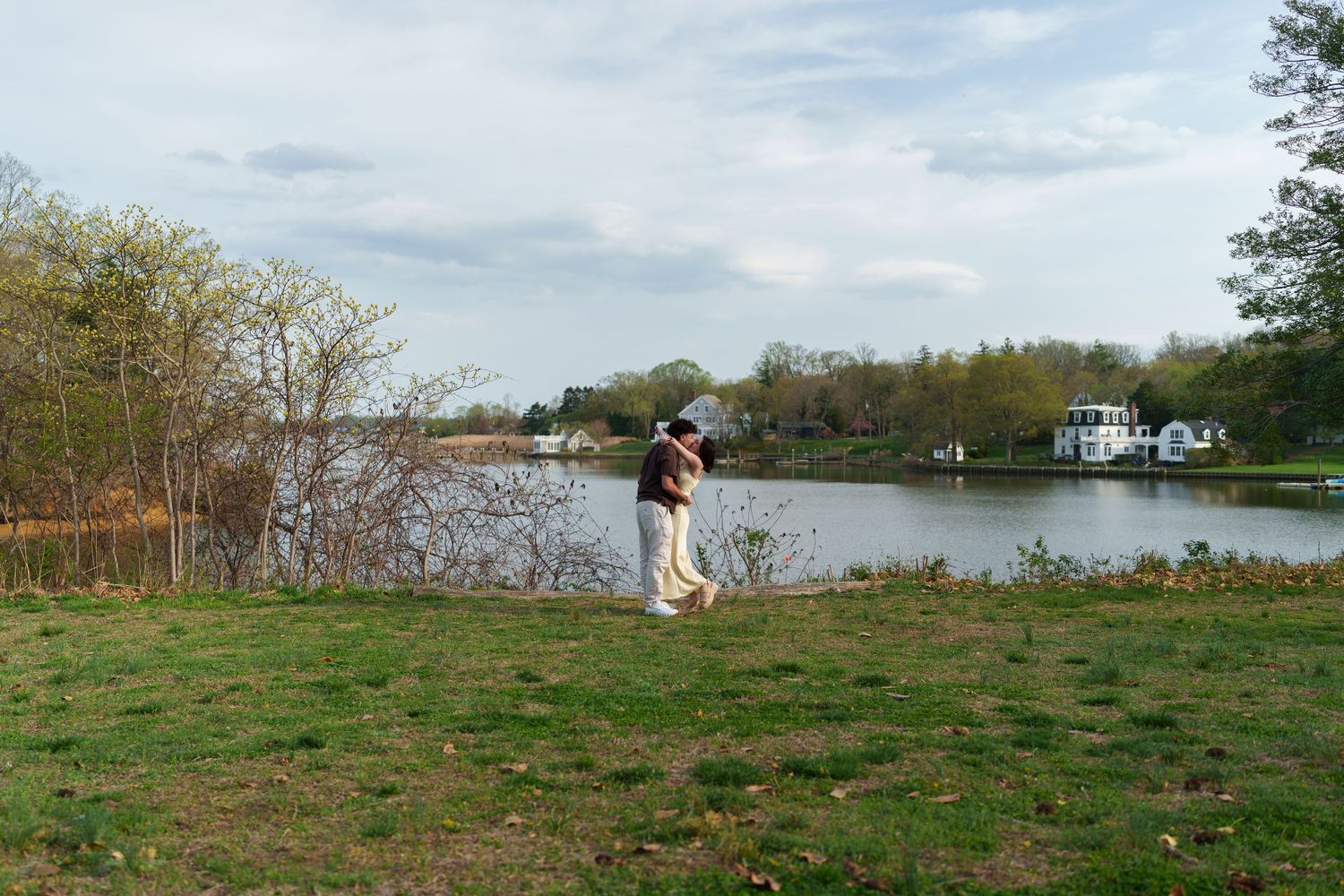 Person standing alone on green grass by a calm river with boats and trees.