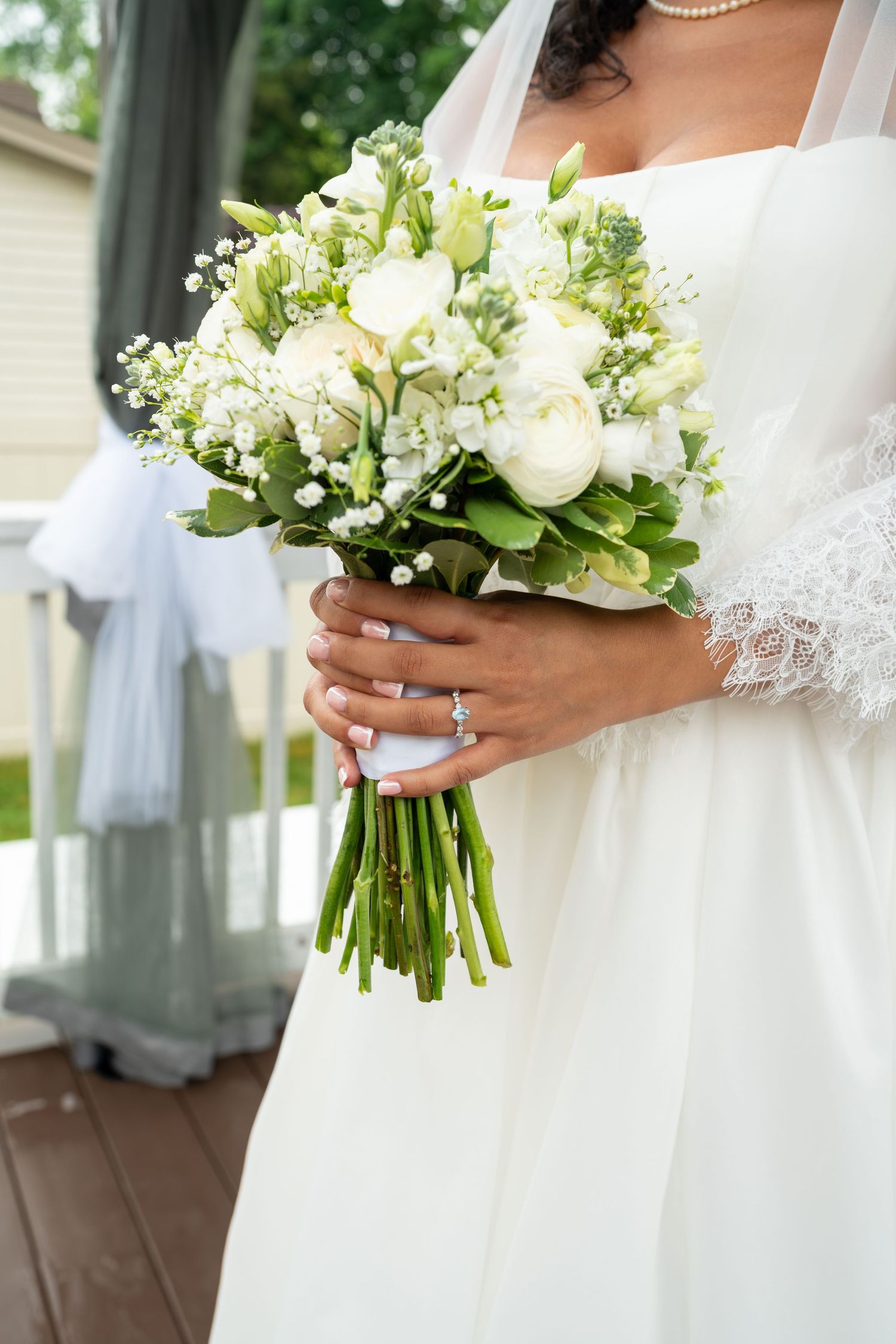 Close-up of a bride holding a lush white rose and greenery bouquet, wearing a diamond engagement ring and pearl bracelet.