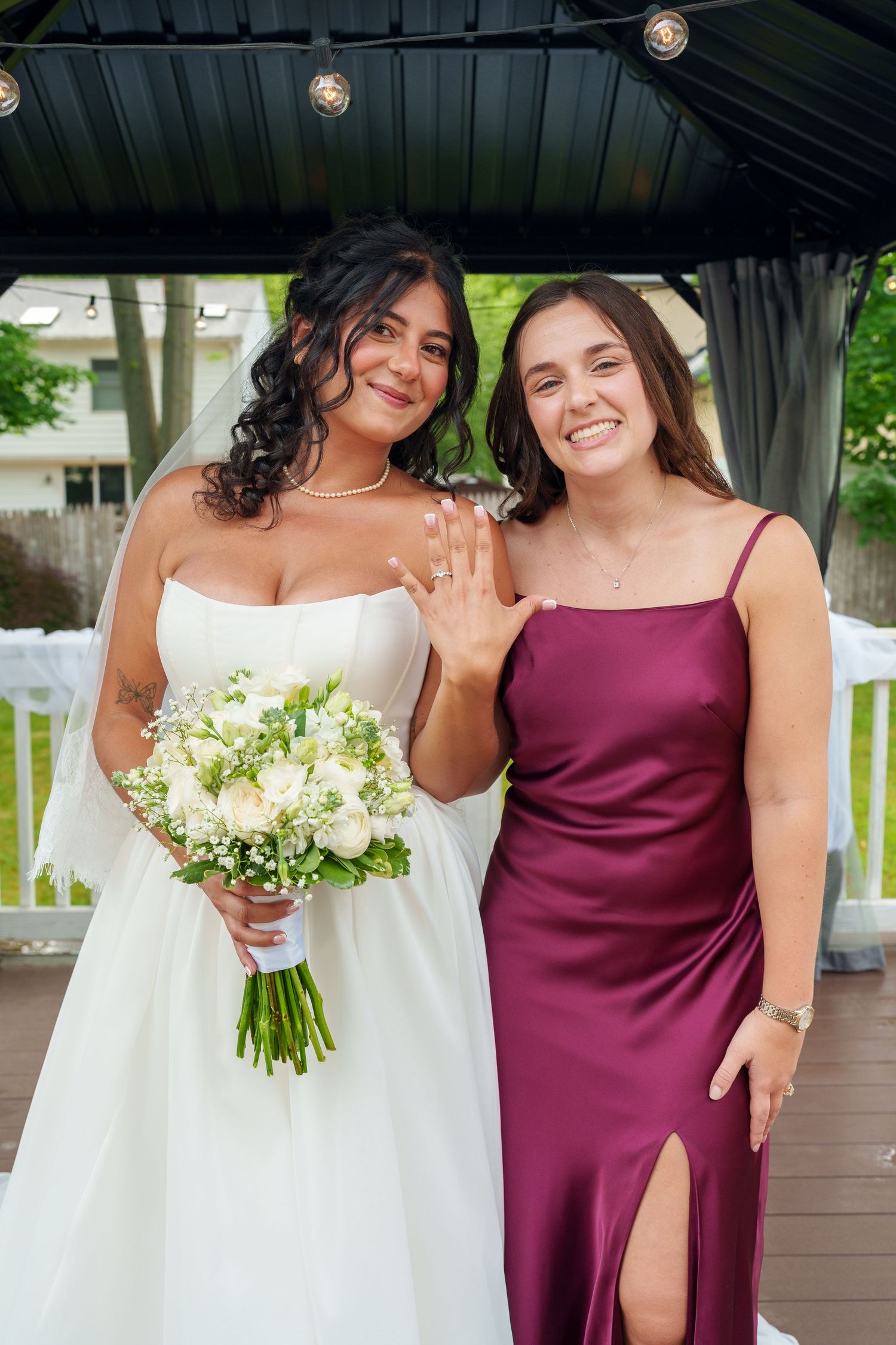 Bride in white gown holding bouquet poses with bridesmaid in burgundy satin dress, showing wedding ring.