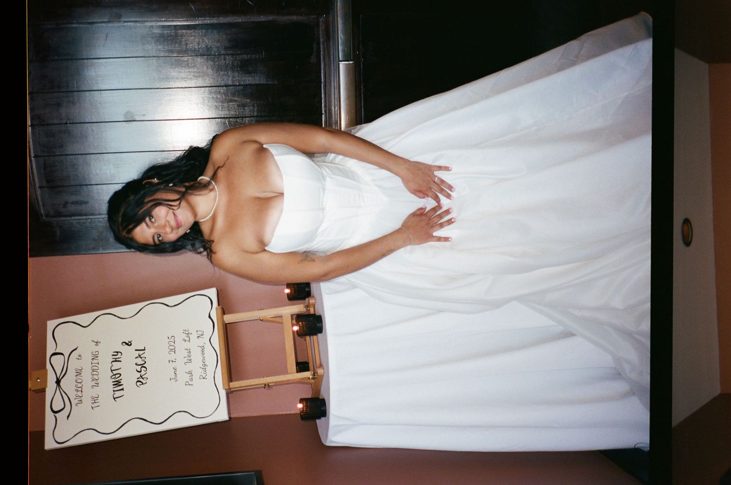 Bride in elegant white wedding gown poses indoors next to a decorative wedding sign display.