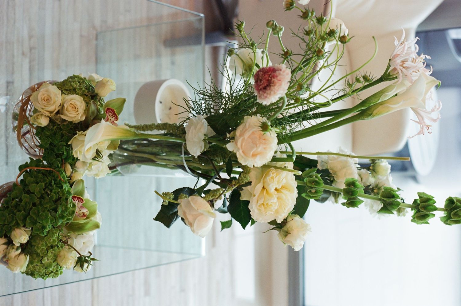 Elegant floral centerpiece with white and blush roses, greenery, and tall stems on a glass surface.