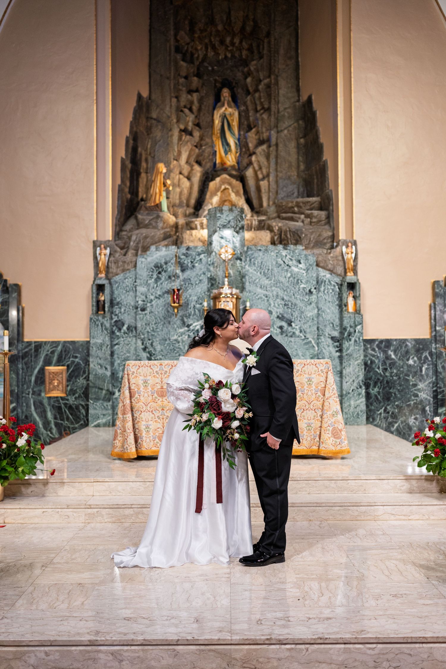 Bride and groom sharing a first kiss at the altar of an ornate Catholic church with elaborate stone backdrop.