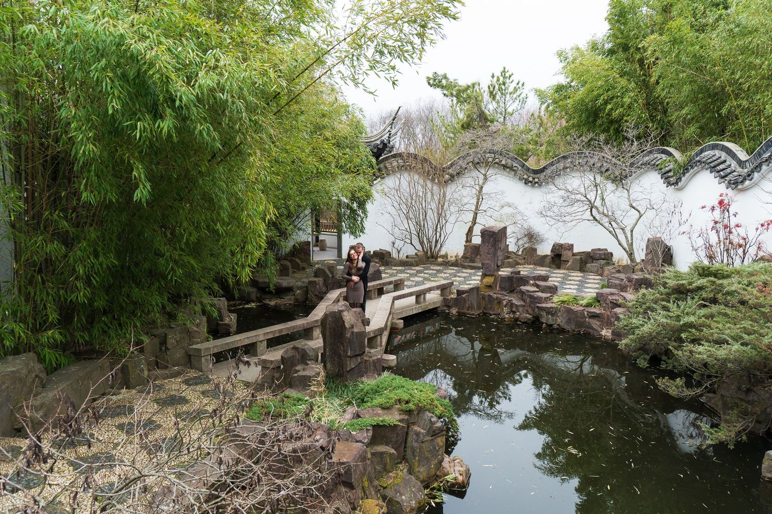 Serene Chinese garden with stone bridge, koi pond, bamboo, and white wall.