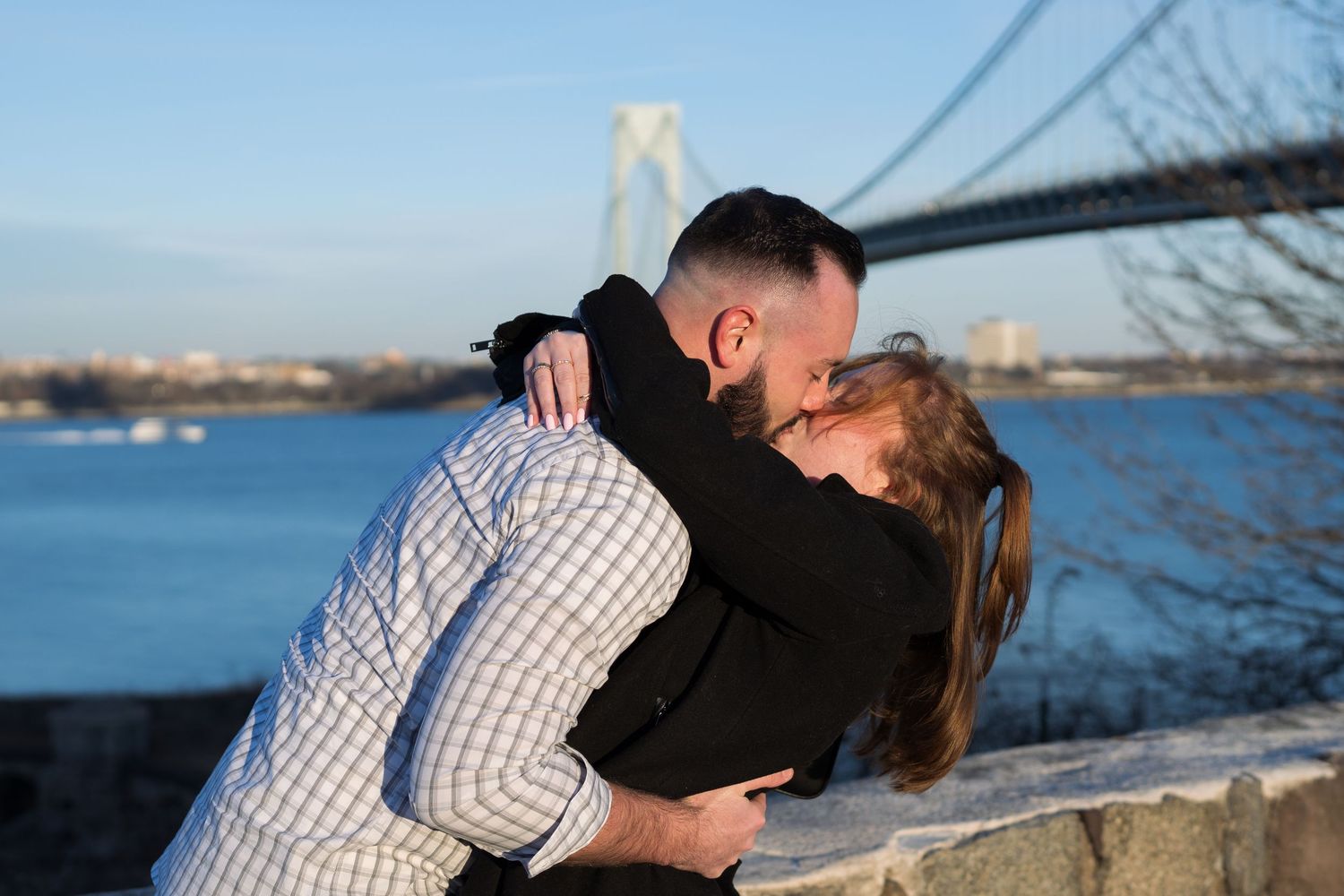 Engaged couple sharing a kiss with the Verrazzano-Narrows Bridge and river visible behind them.
