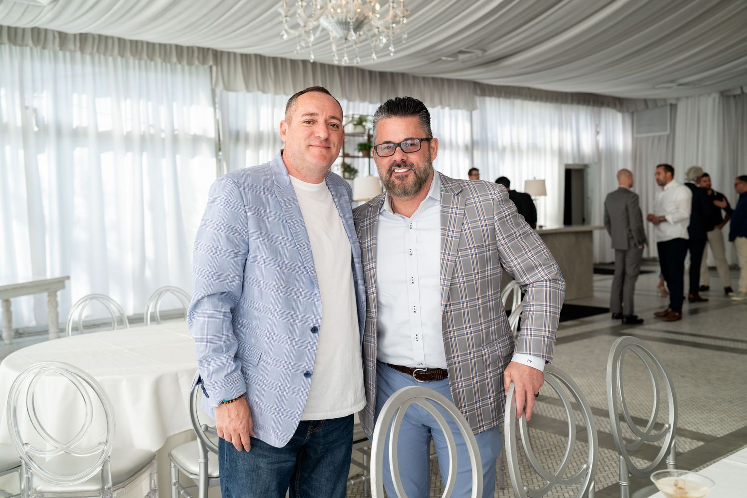 Two men in blazers posing together at an elegant white-draped event venue with ghost chairs.