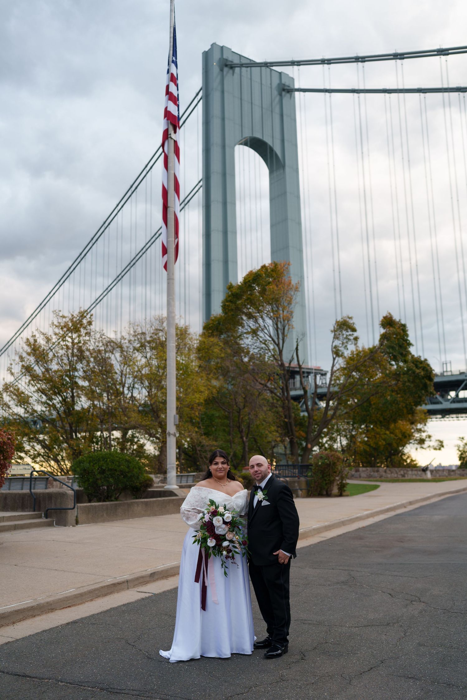 A bride and groom stand together near an American flag with the Verrazzano-Narrows Bridge as a backdrop.