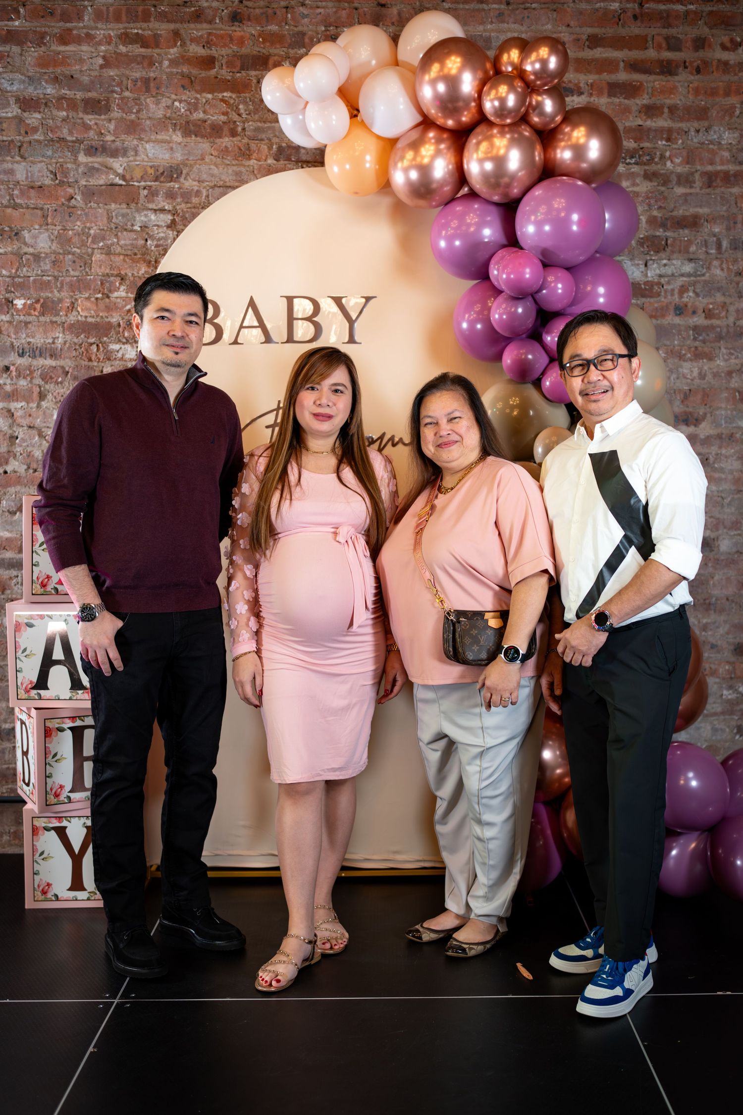 Pregnant woman in pink dress poses with family at baby shower balloon arch backdrop.