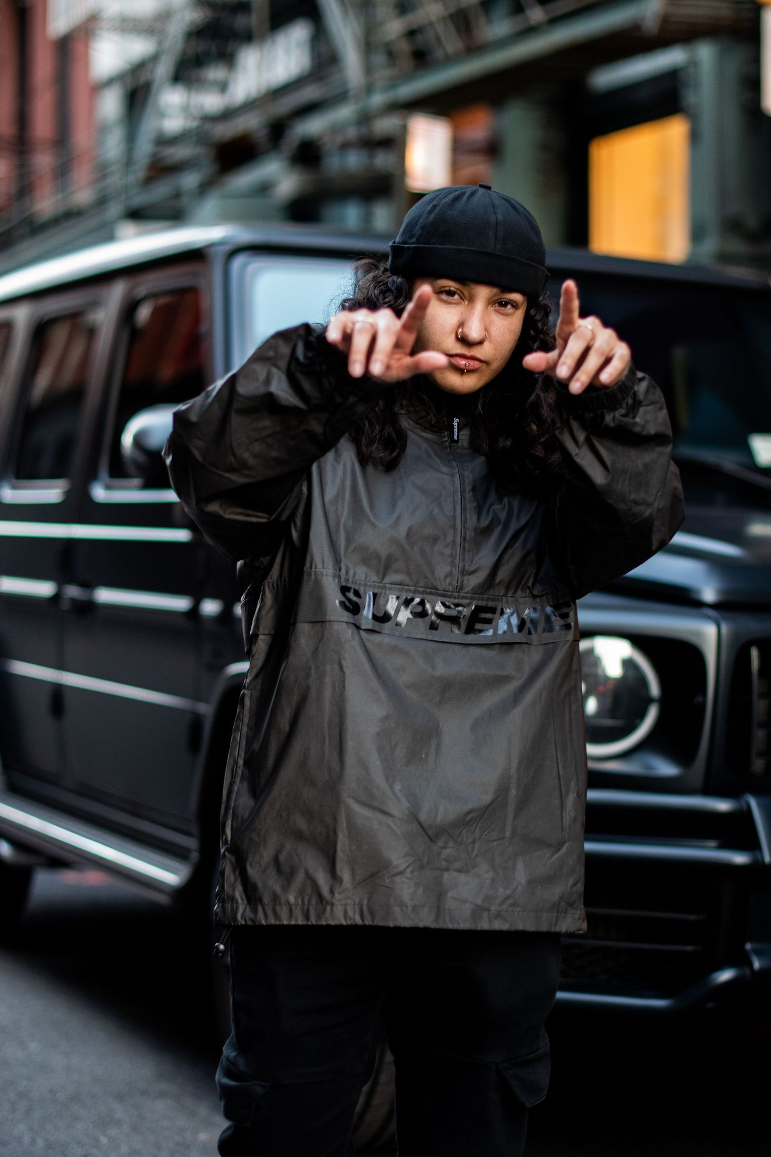 A person in black streetwear poses in front of a black Mercedes G-Wagon on a city street.