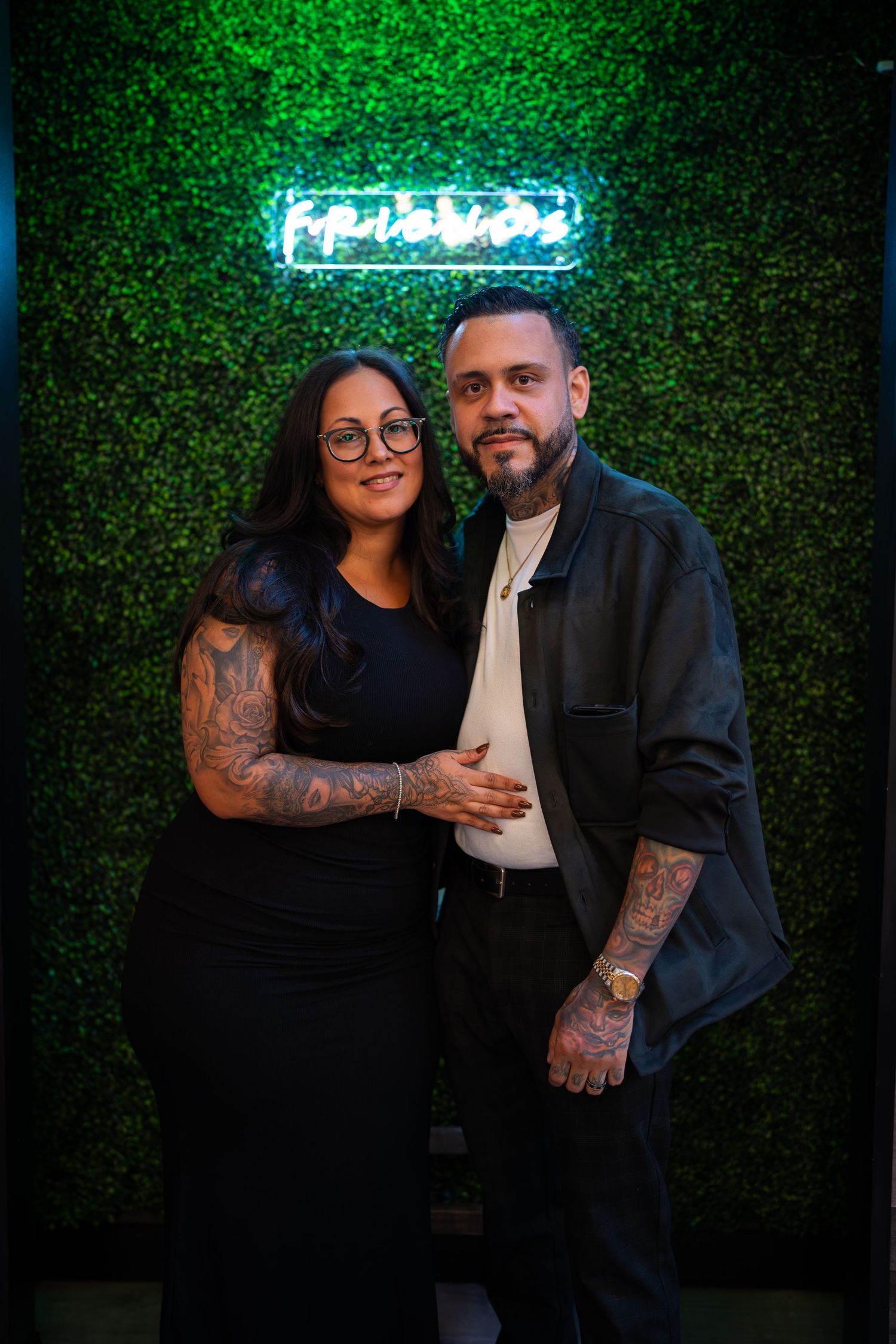 Tattooed couple posing together in front of a green hedge wall with neon sign.