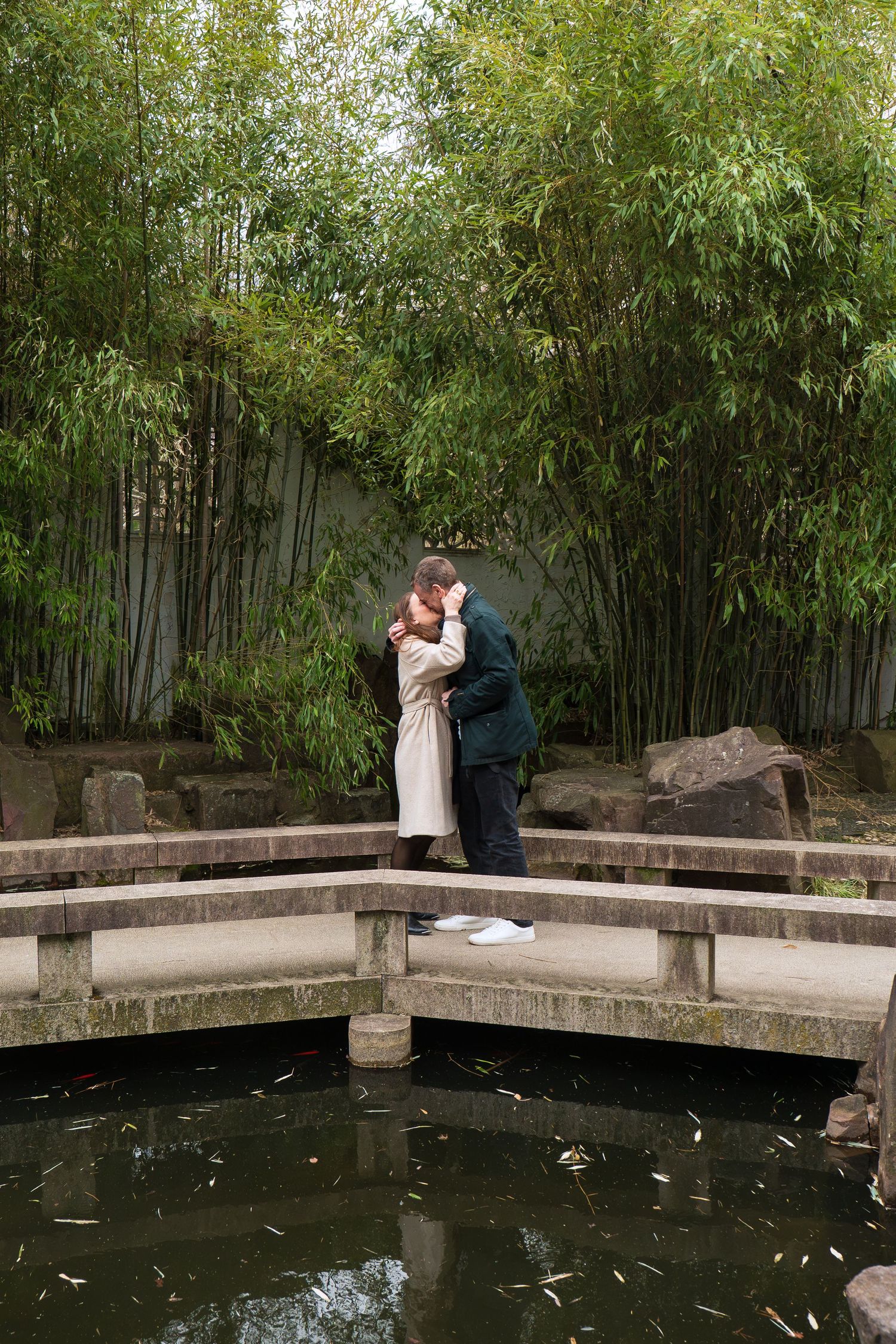 Couple sharing a romantic kiss on a curved stone bridge over a serene pond in a bamboo garden.