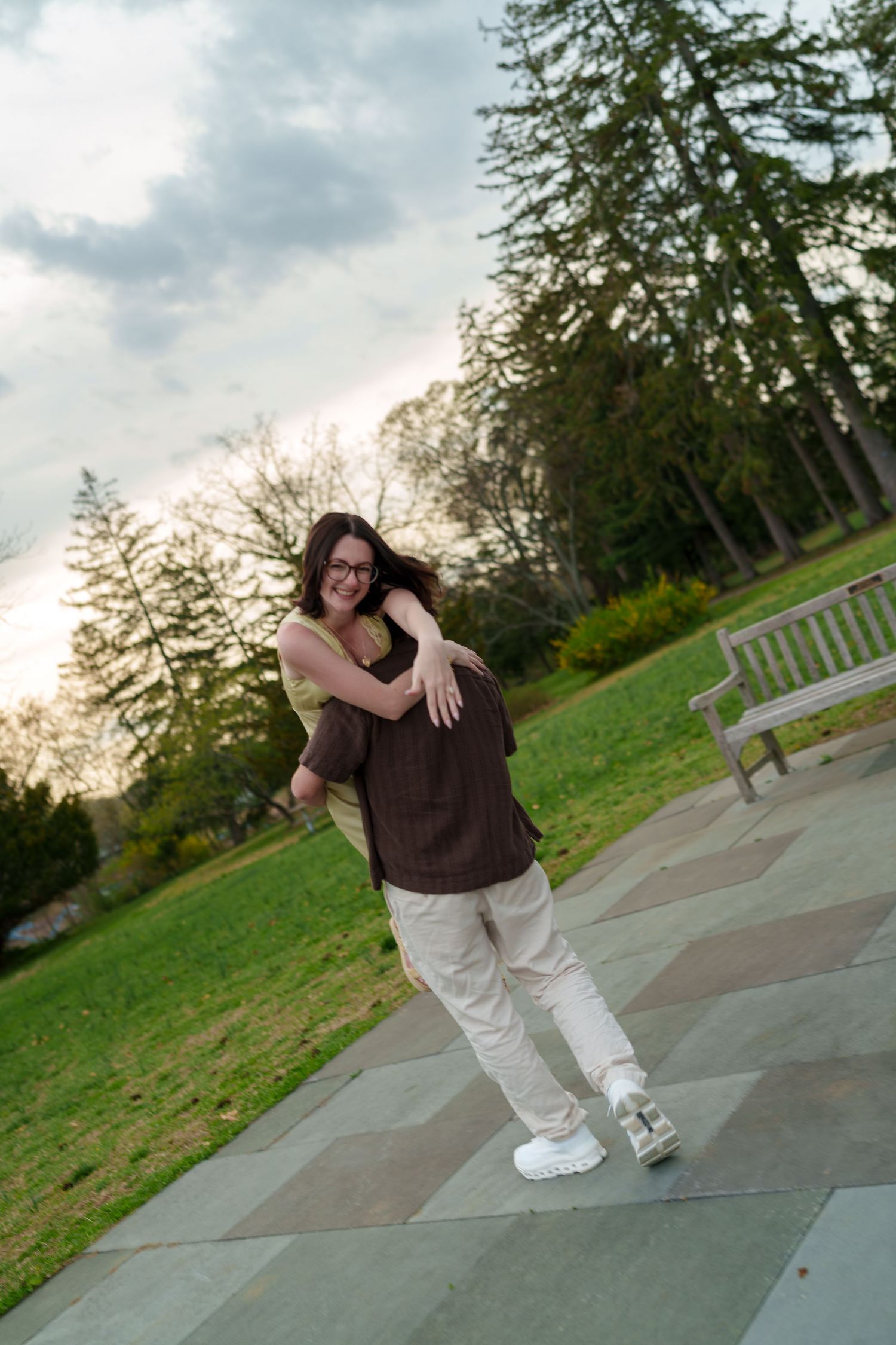 Woman hugging man from behind on a scenic park pathway at sunset with tall trees nearby.