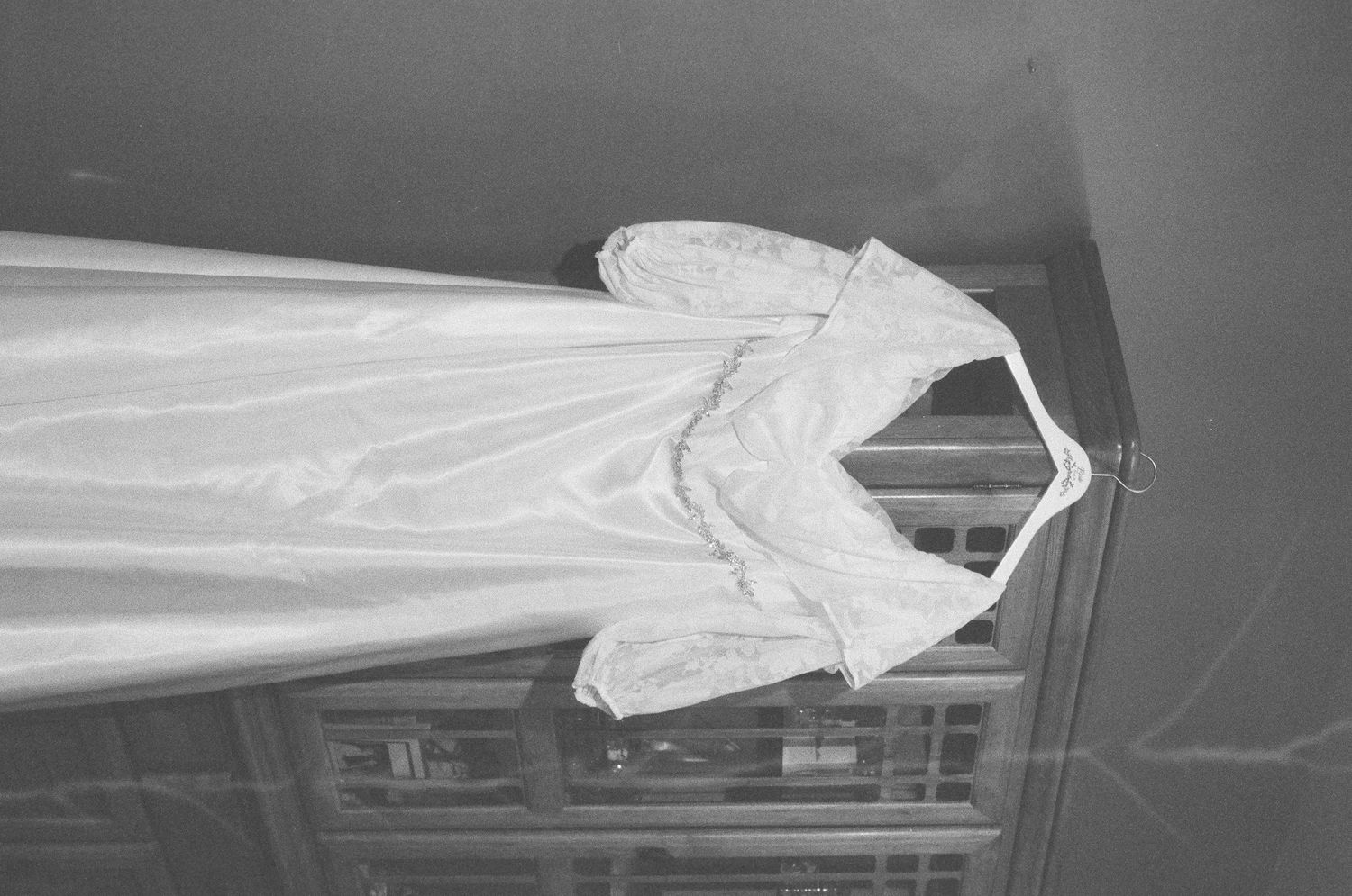 Black and white photograph of a beautiful white wedding dress hanging gracefully near a window.