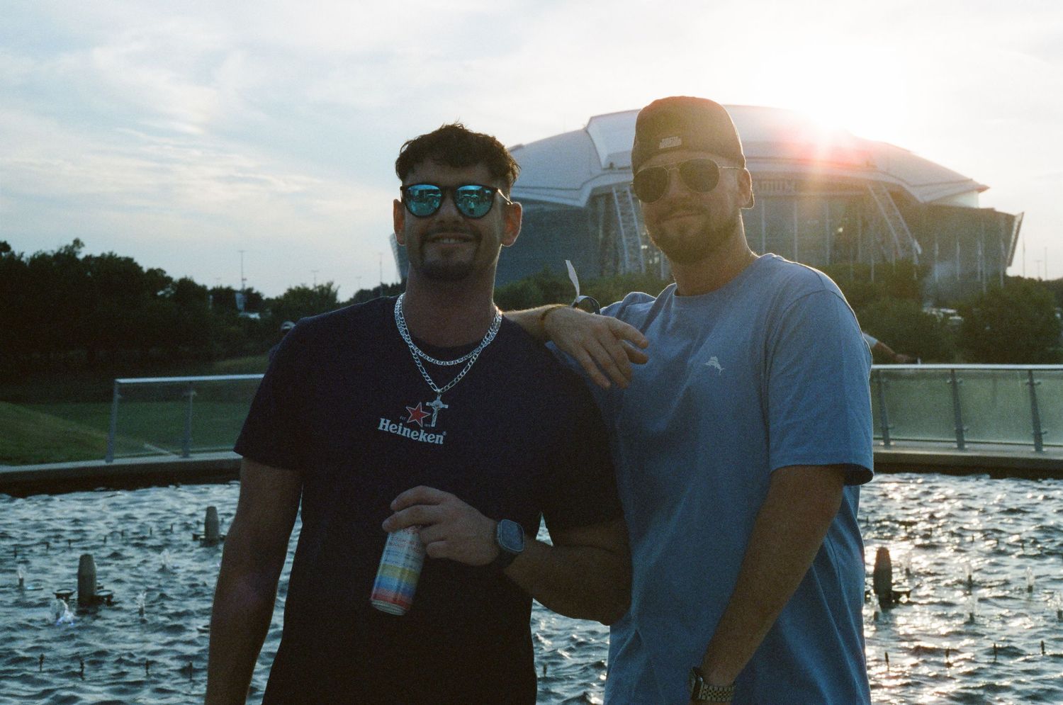 Two young men pose and smile in front of a large stadium at sunset near a reflecting pool.