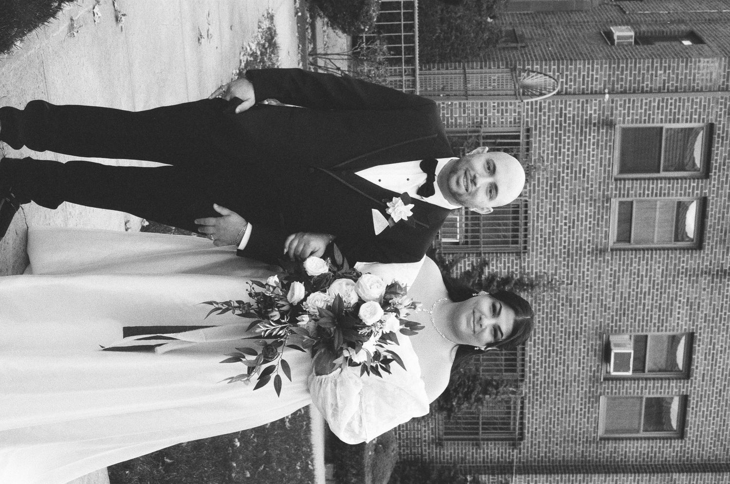 Black and white photo of a bride and groom posing together outdoors, bride holding a floral bouquet.