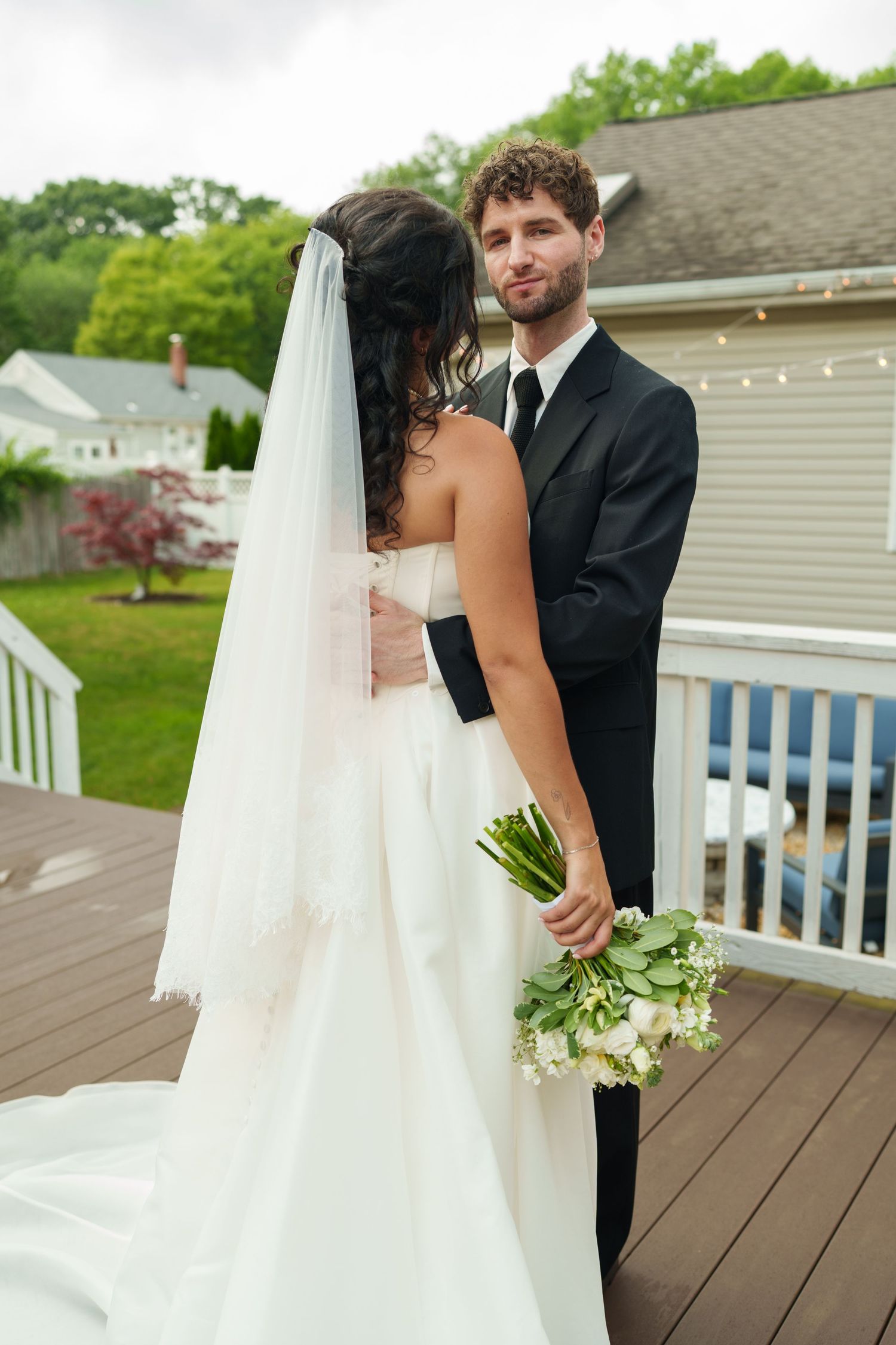 Groom in black suit holding bouquet gazes at camera while embracing bride in white gown with veil on outdoor deck.