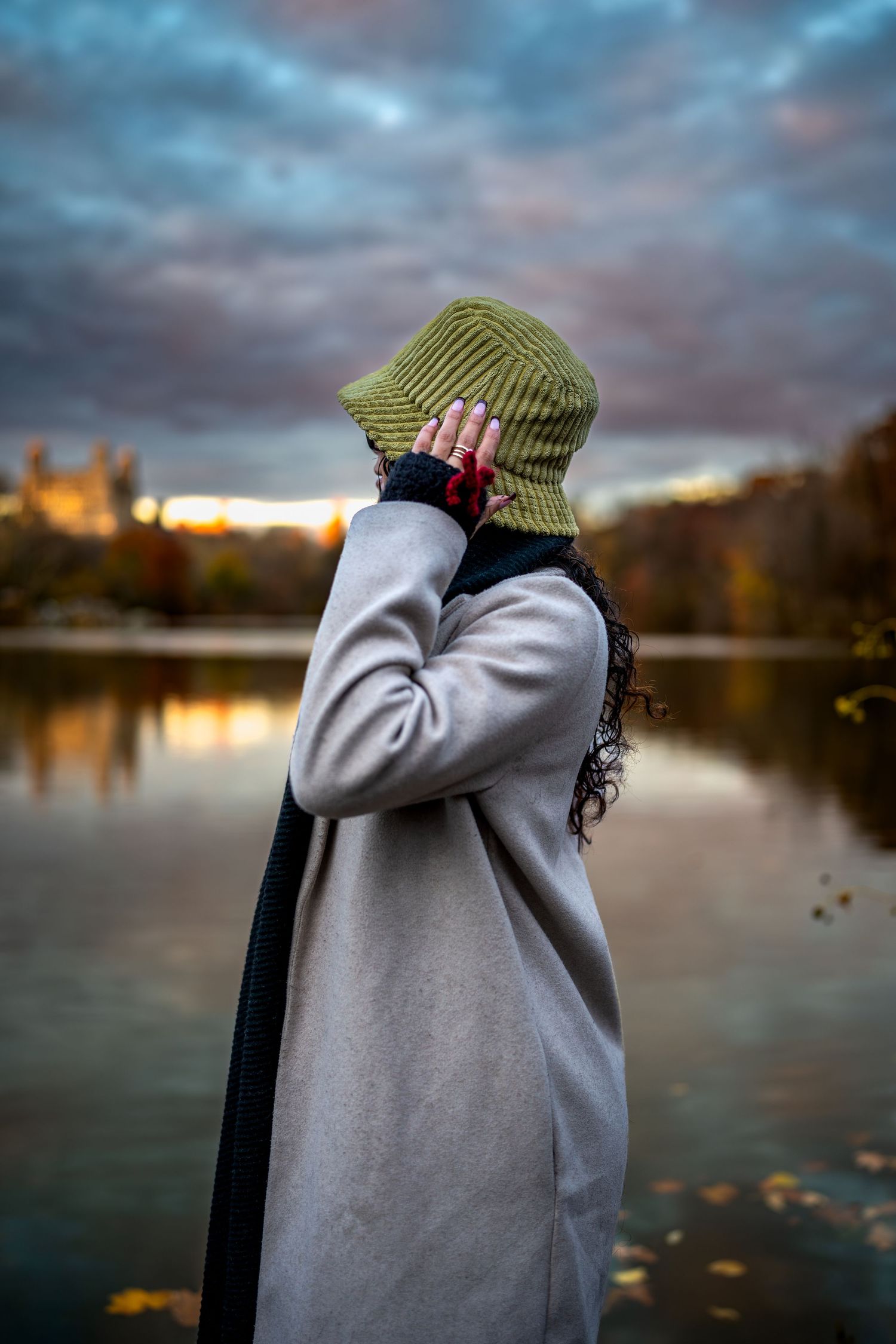 Woman in gray coat covers face with green bucket hat near reflective autumn lake at dusk.