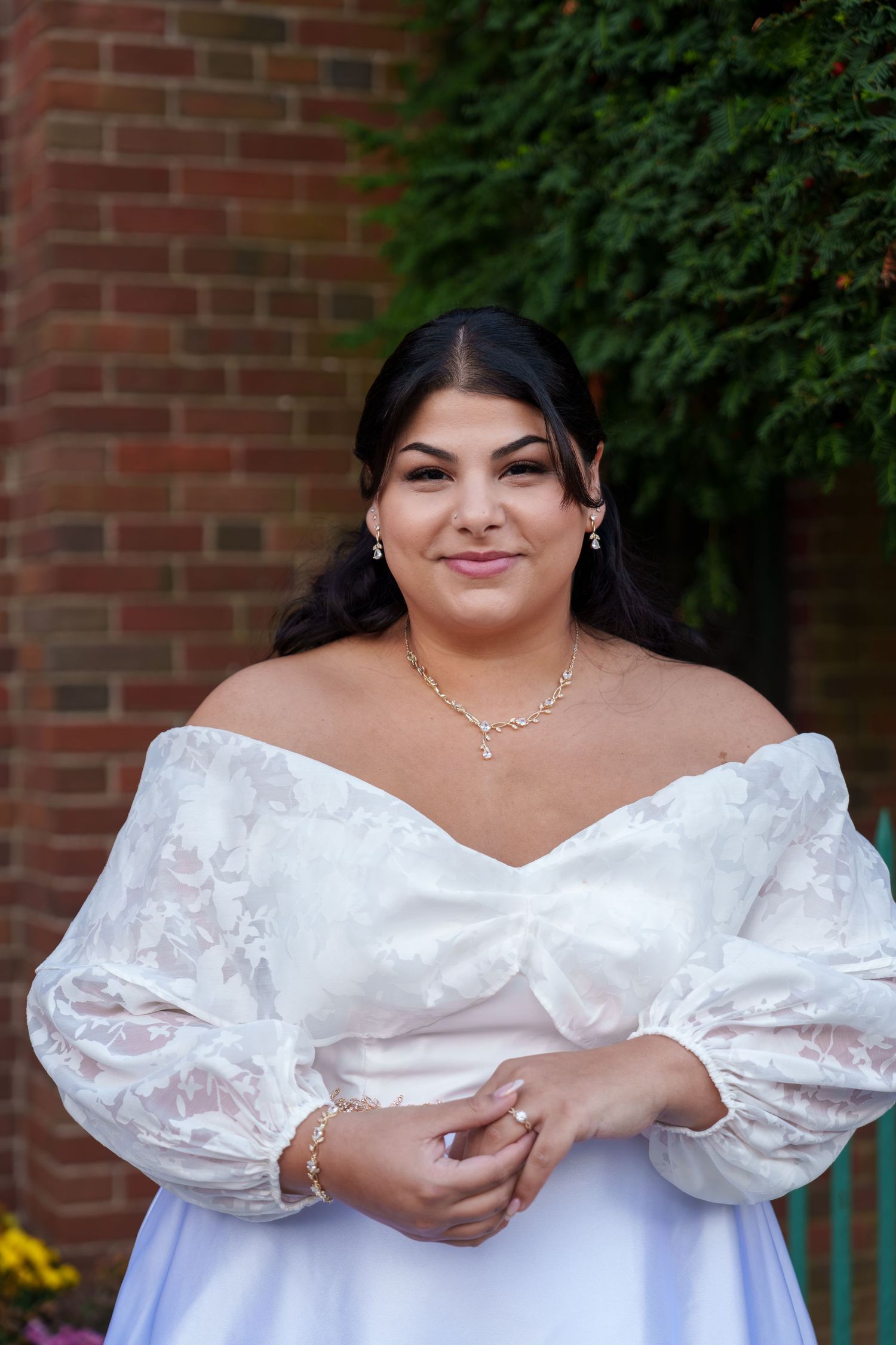 A beautiful bride in an off-shoulder white lace wedding gown smiles outdoors at her wedding.