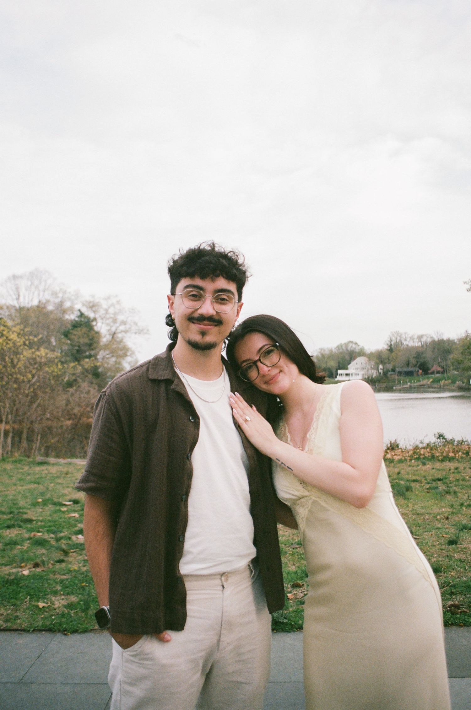 Couple smiling together outdoors near a lake, woman in yellow dress leaning on mans shoulder.