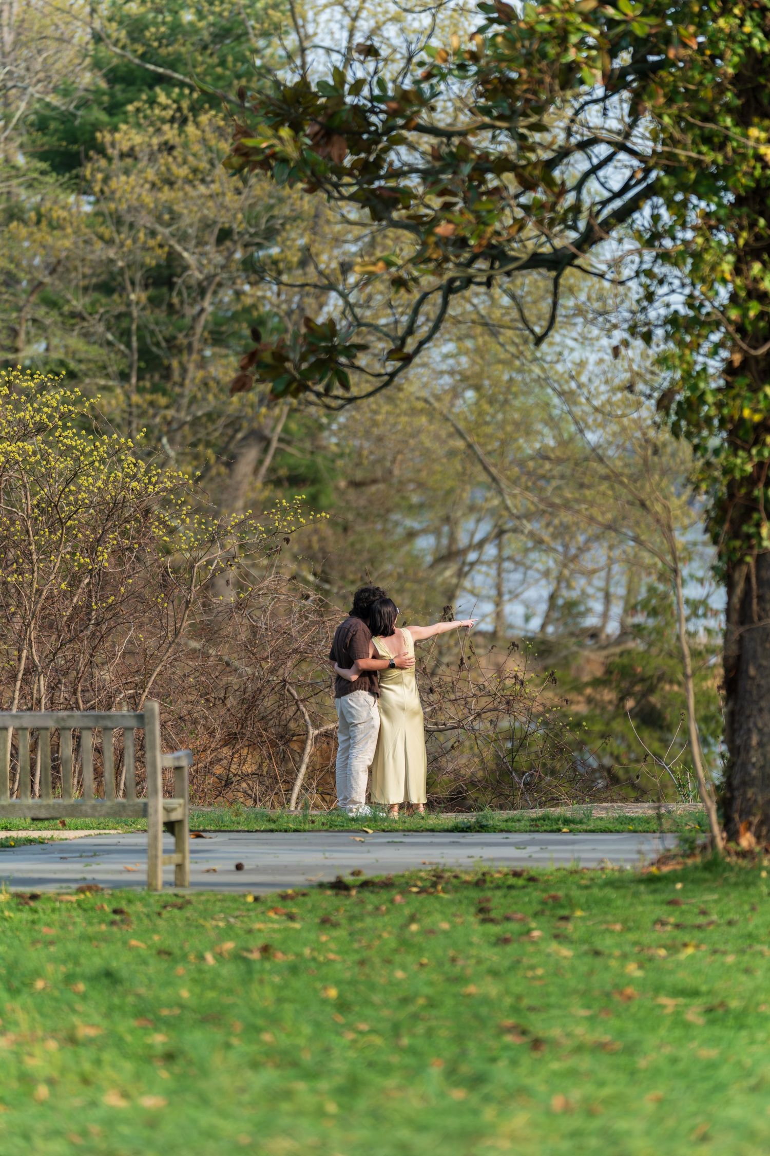Couple embracing on a park path, one pointing into the distance near a wooden bench.