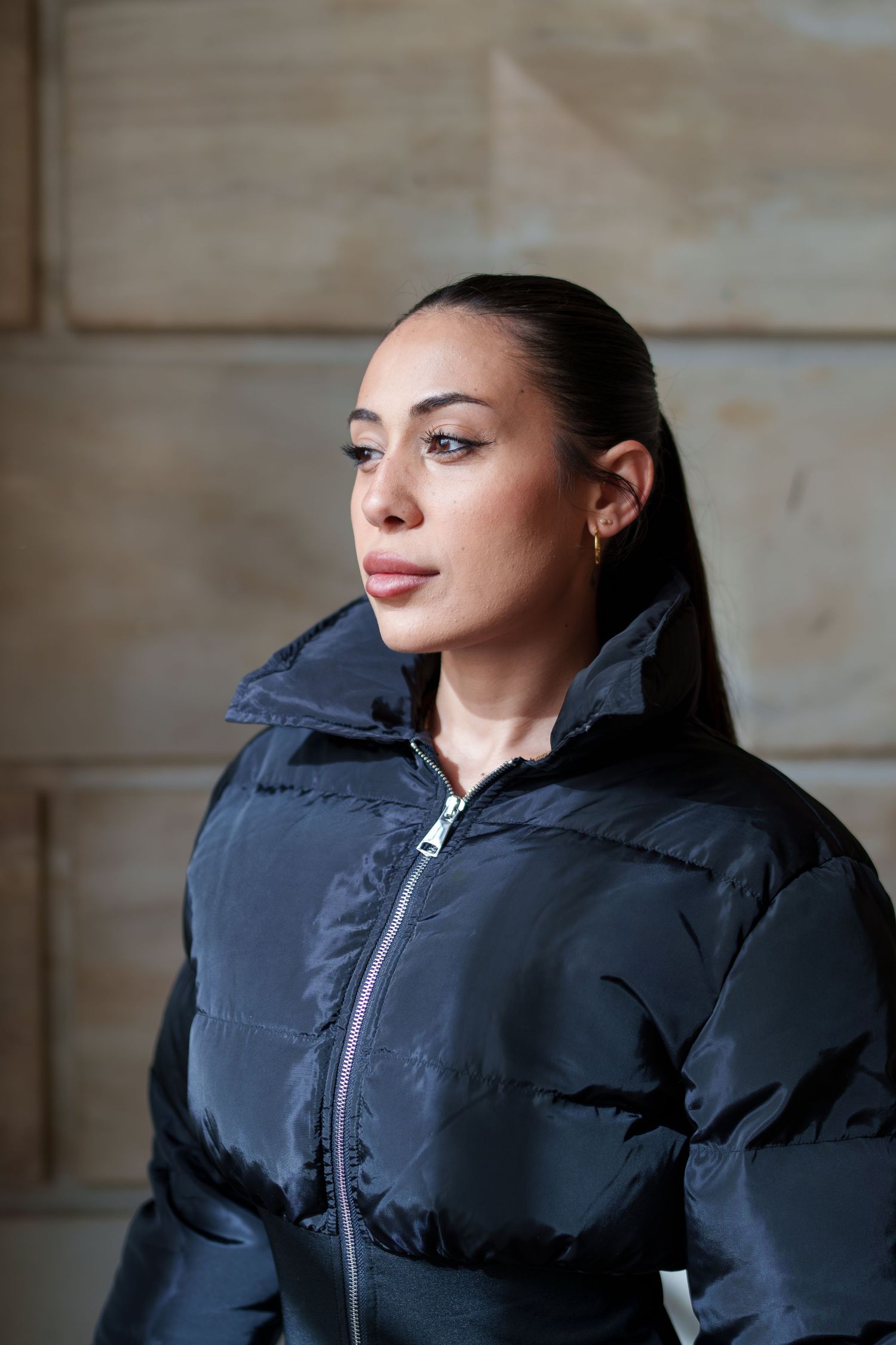 Close-up portrait of woman in black puffer jacket with sleek ponytail against stone wall.