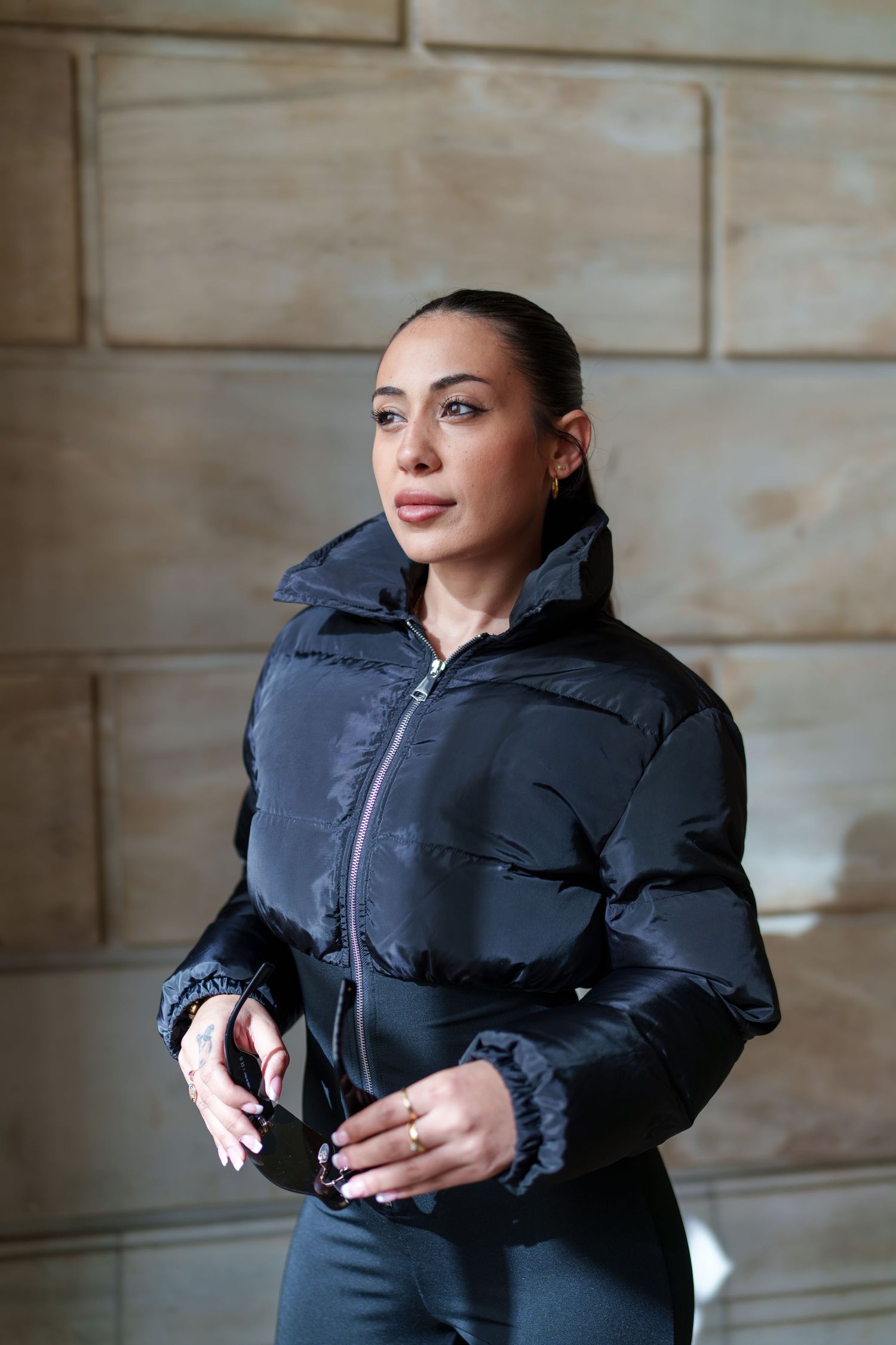 Woman in black zip-up cropped puffer jacket posing confidently against stone wall background.