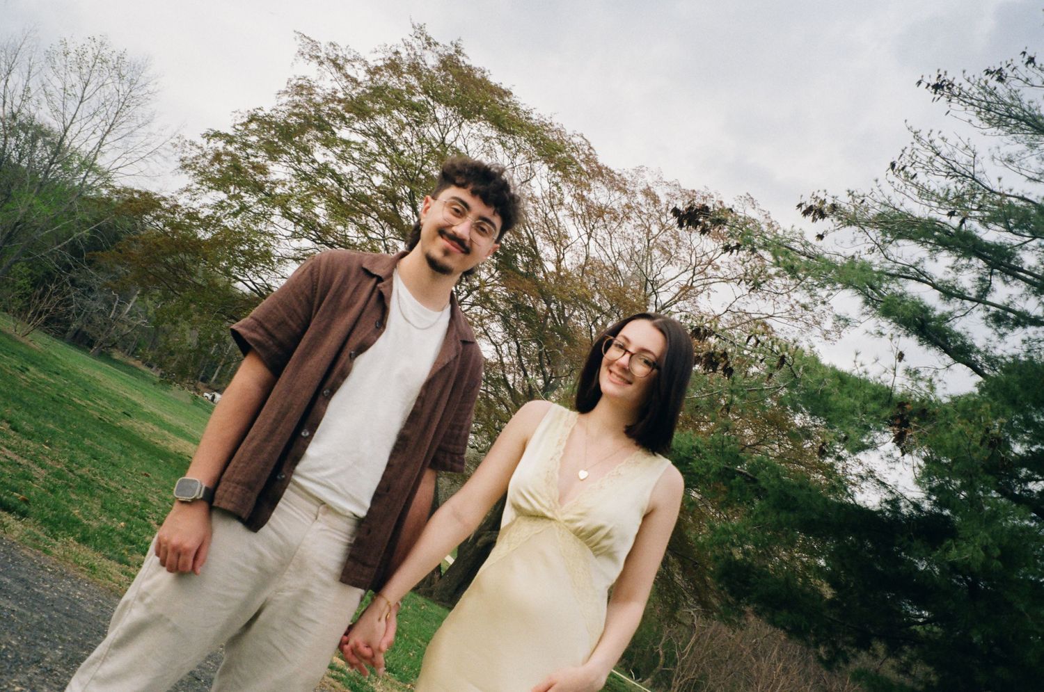 Couple holding hands and smiling in a lush green park with blooming trees.
