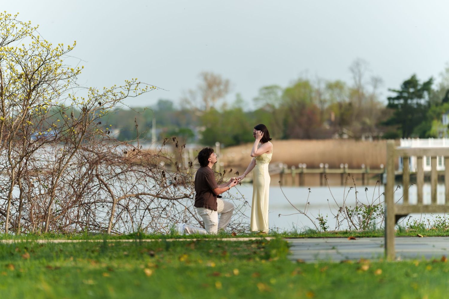 Man kneeling to propose to woman in yellow dress by a scenic waterfront boardwalk.