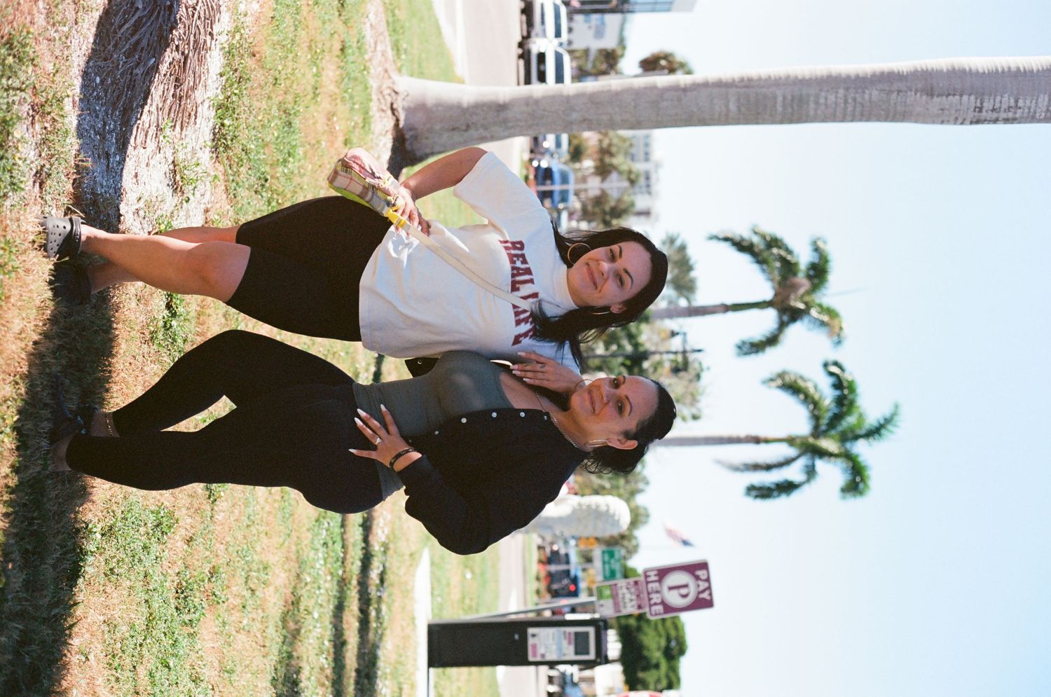 Two young women posing and leaning against a palm tree in a sunny outdoor park setting.