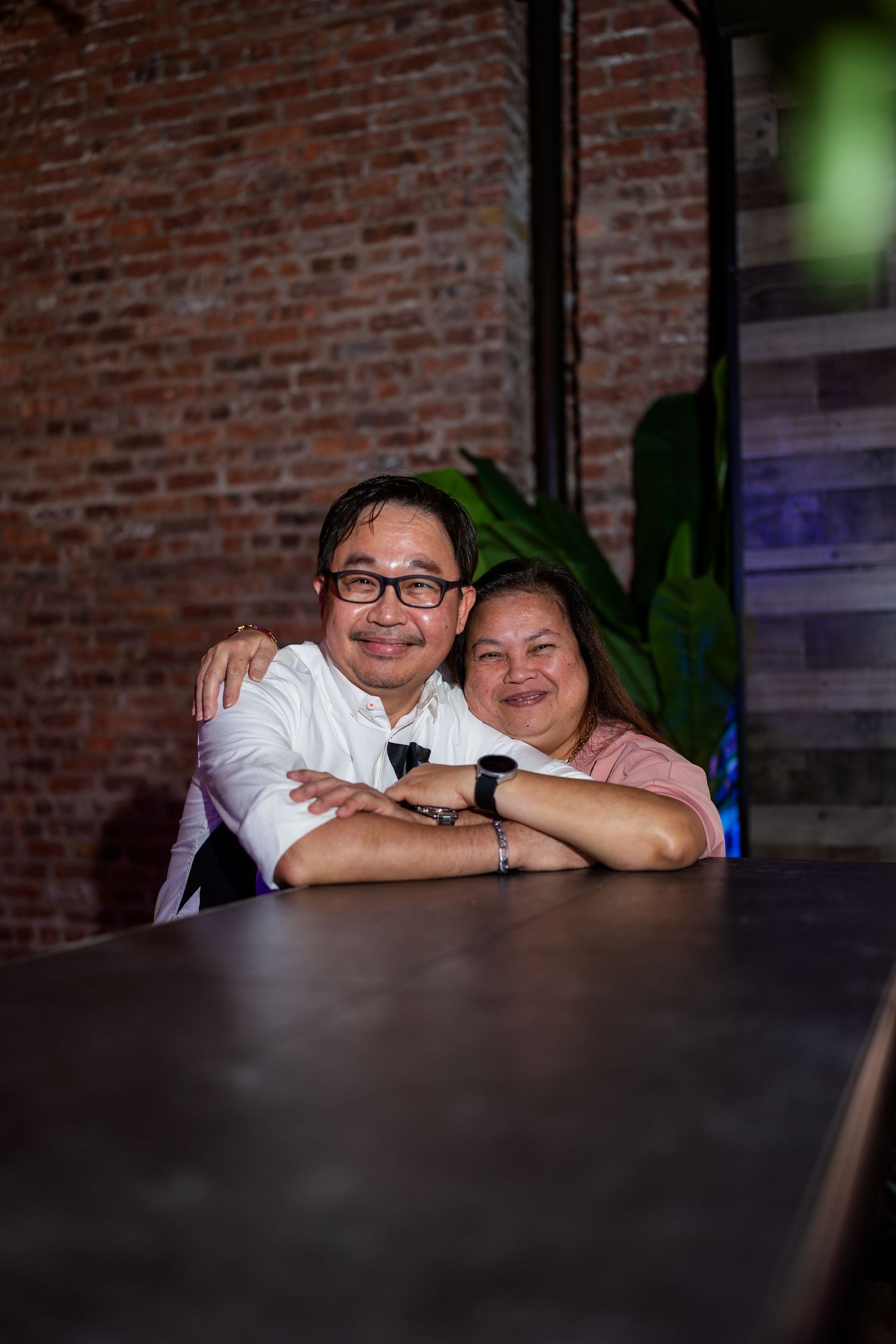 A smiling couple embracing warmly while seated at a table in front of a brick wall.
