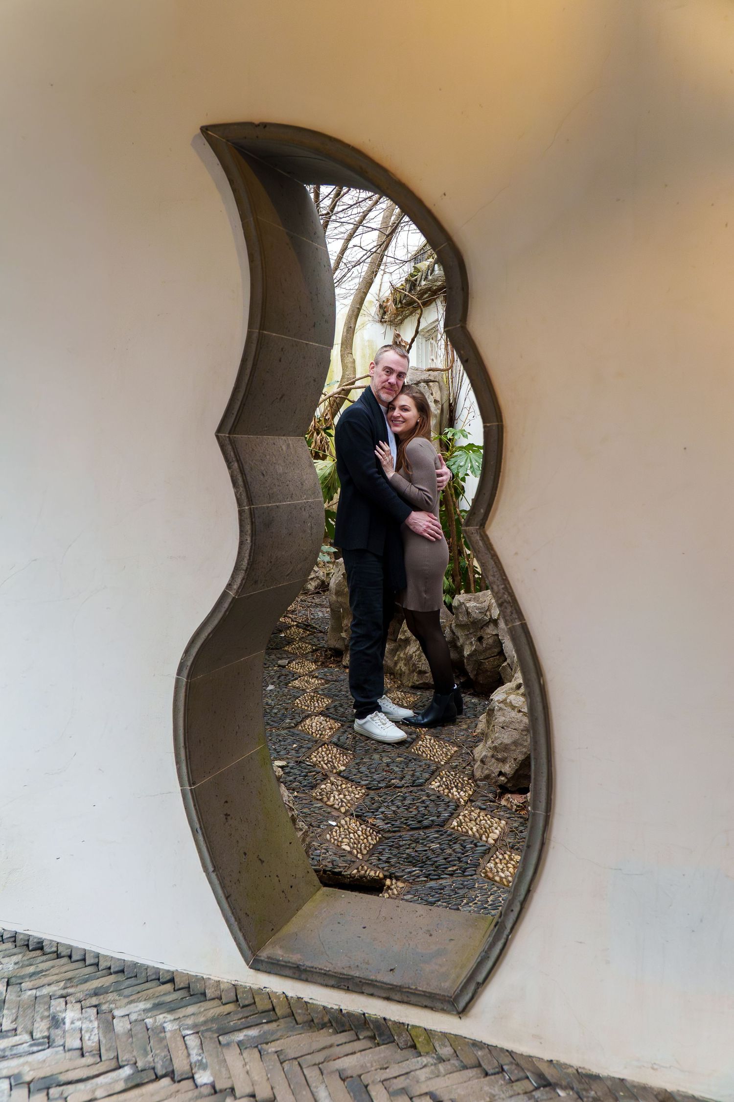 Couple embracing framed by an ornate flame-shaped moon gate in a Chinese garden wall.