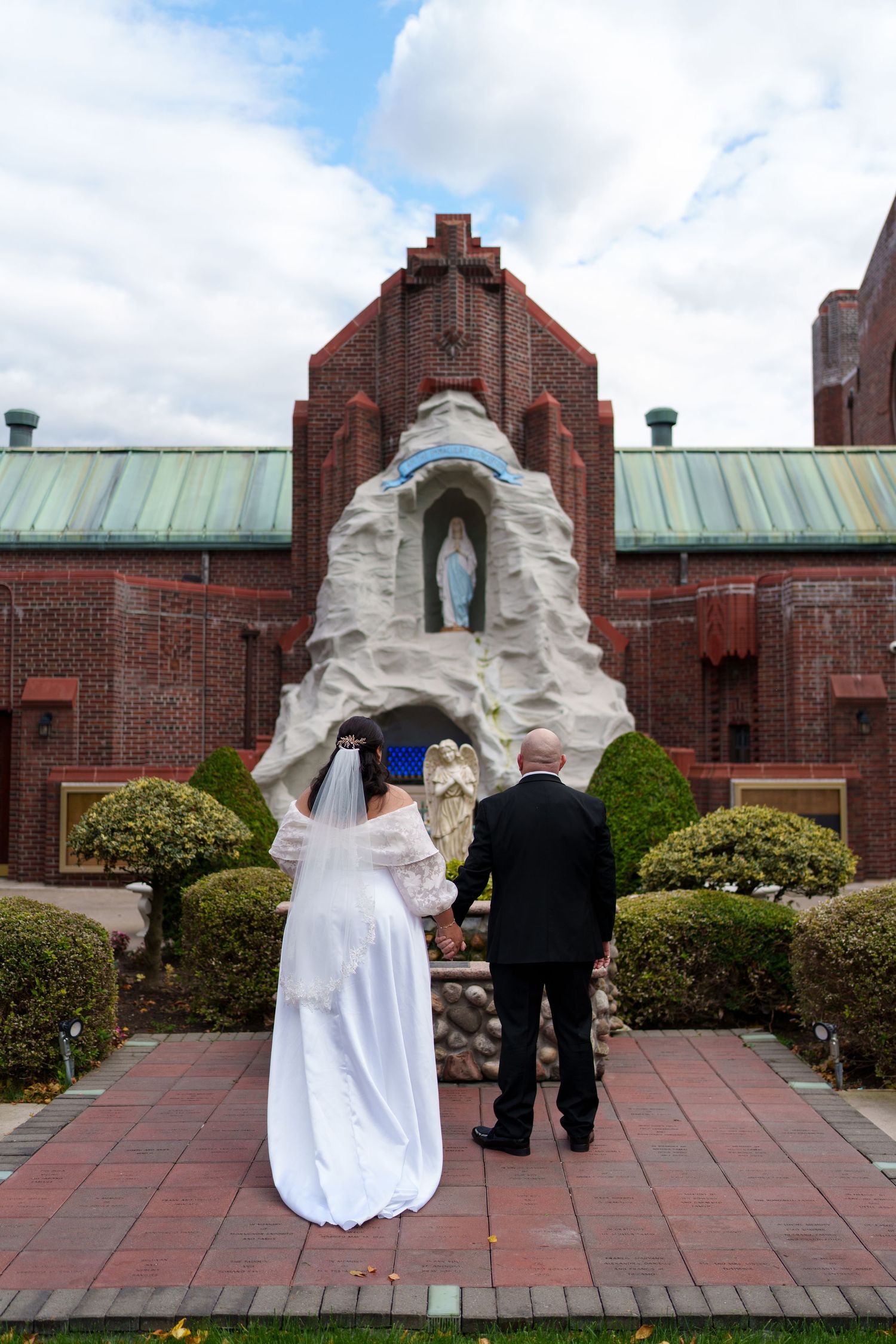 A bride and groom hold hands while facing a religious grotto statue outside a red brick Catholic church.