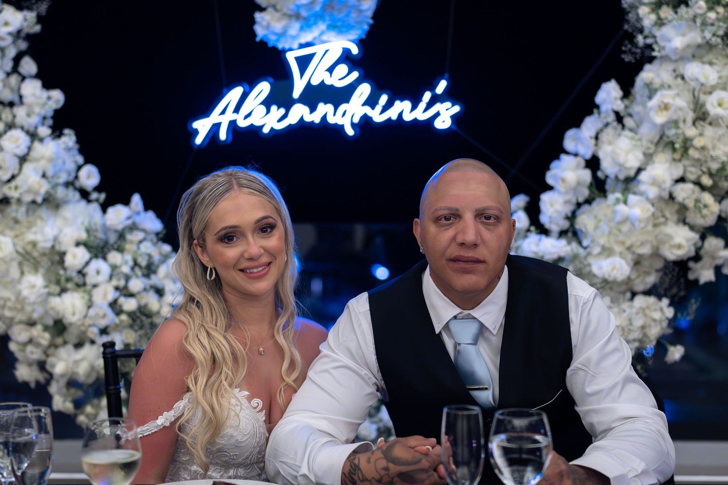 Newlyweds sit at reception table beneath glowing neon sign reading The Alexandrini's surrounded by white florals.