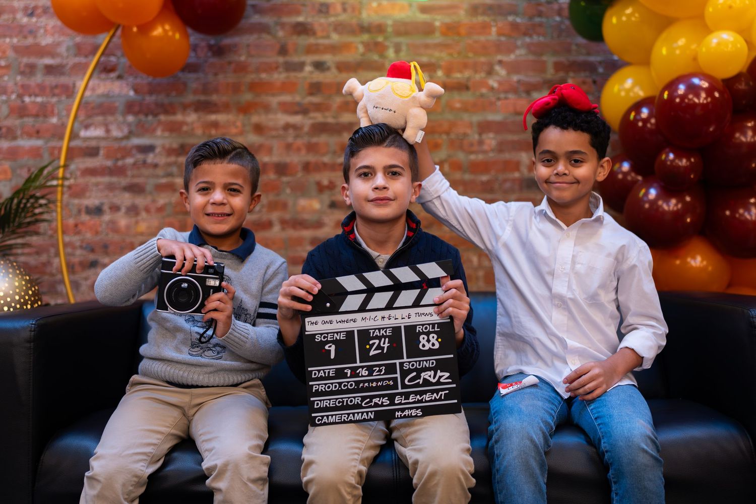 Three young boys posing with a film clapperboard and stuffed animal at a party.