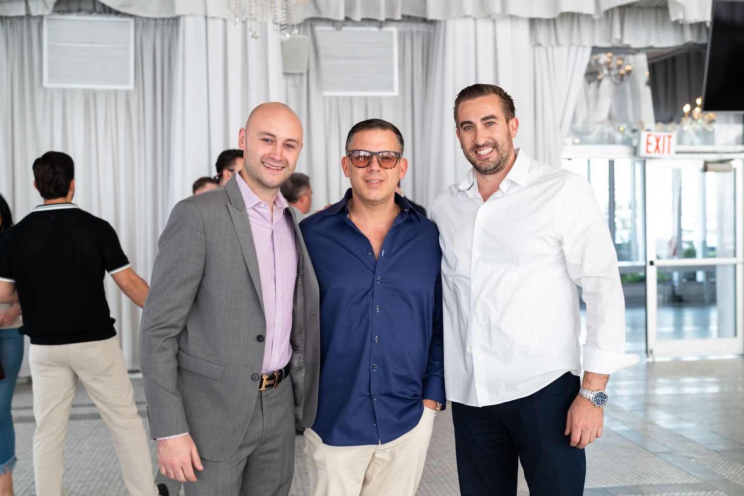 Three smiling men in business casual attire posing together at an indoor networking event.