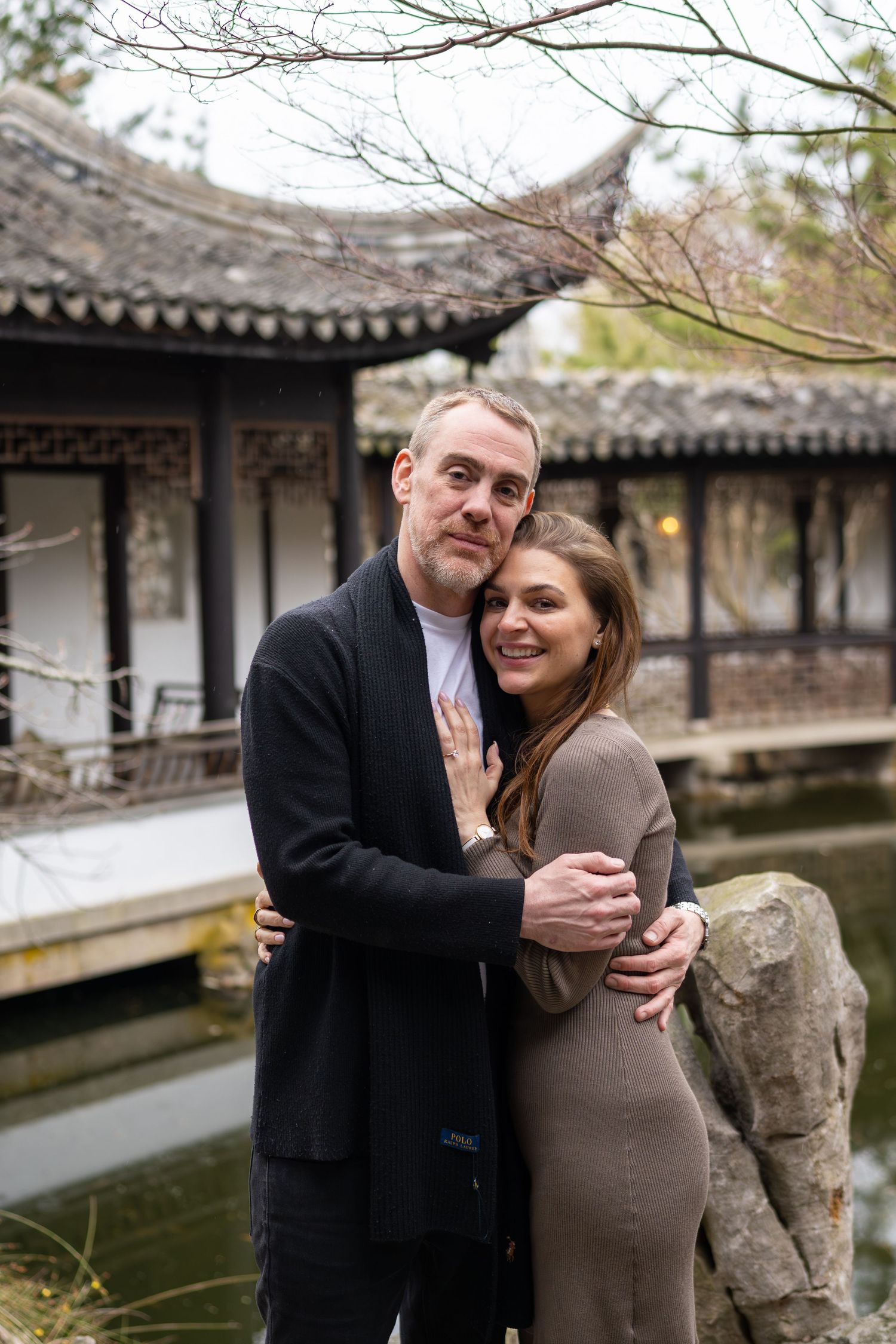 Smiling engaged couple embracing by a reflective pond with a classic Chinese pavilion behind them.