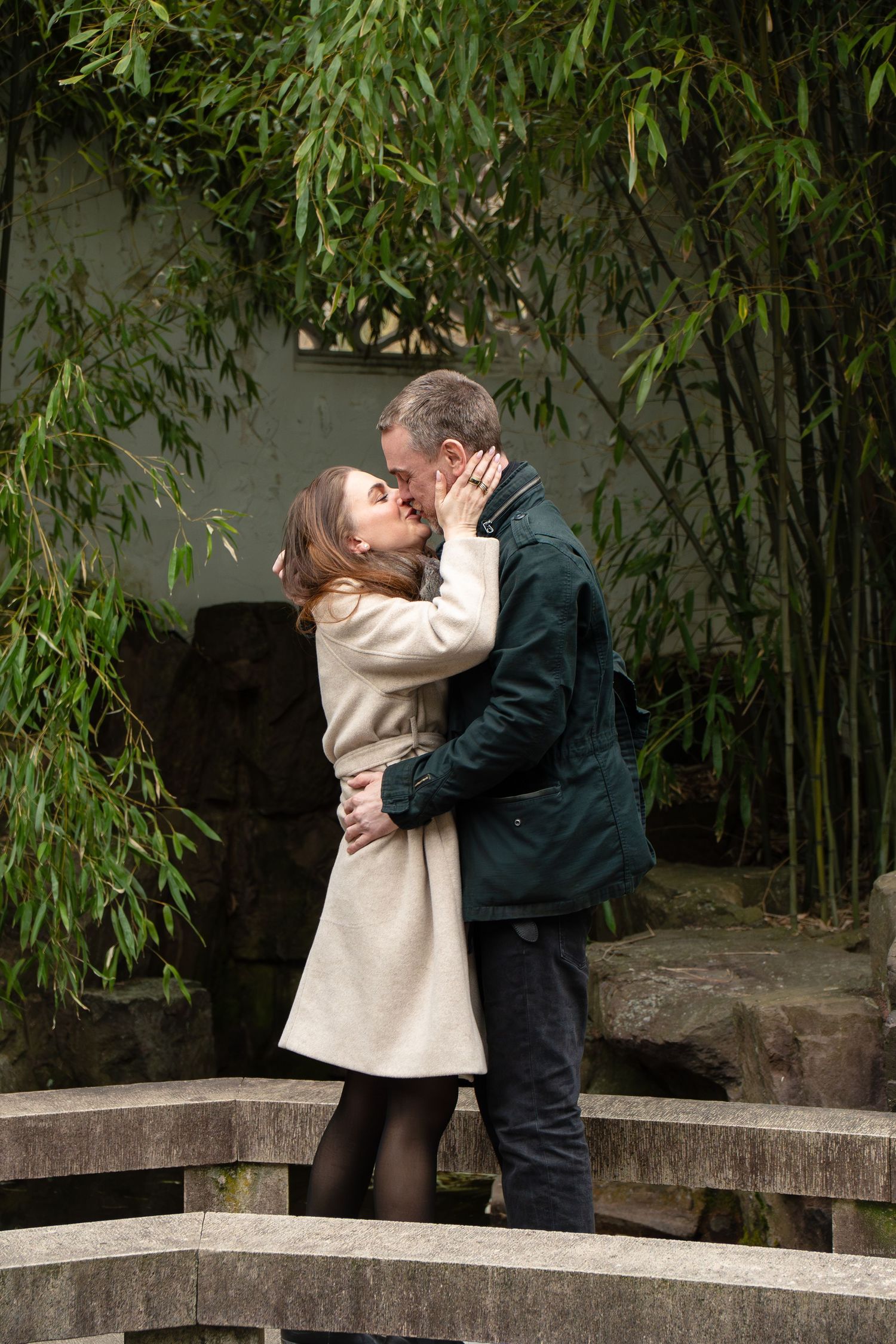 Couple kissing passionately on a stone bridge surrounded by tall green bamboo in a tranquil garden.