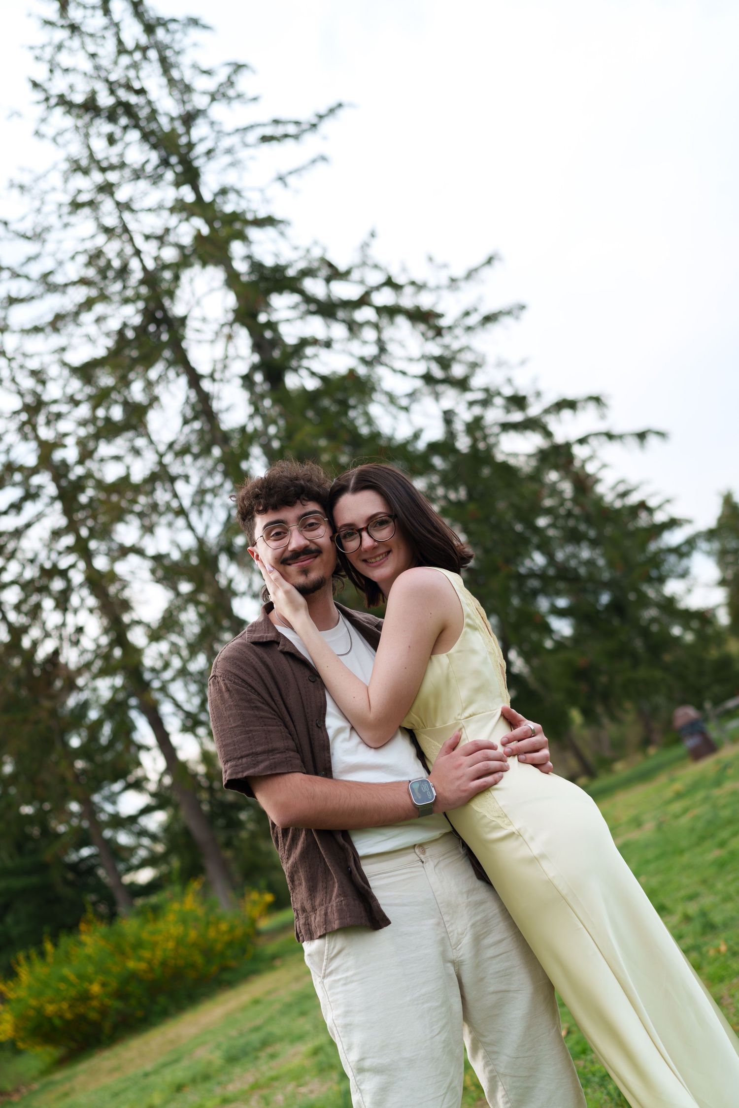 Happy couple embracing and smiling in a park with tall evergreen trees in background.