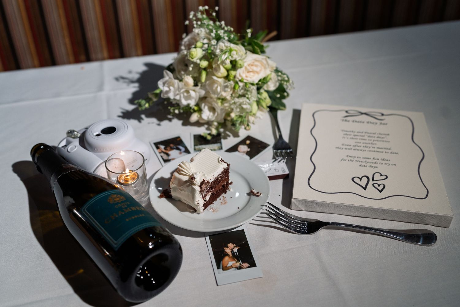Wedding sweetheart table with champagne bottle, cake slice, polaroids, white floral centerpiece, and menu card.