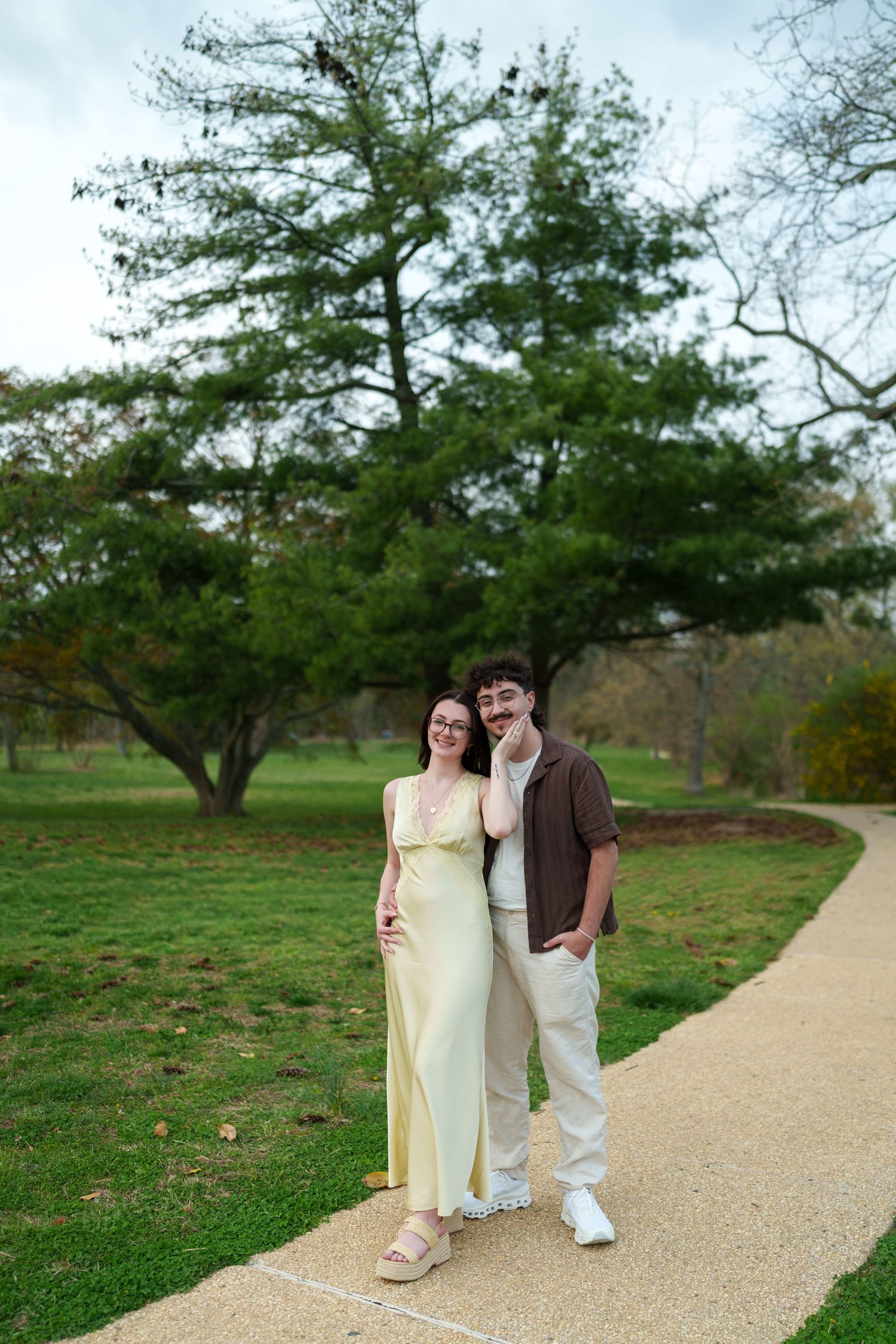 Couple standing together and smiling on a park pathway, woman in yellow maxi dress and man in brown shirt.