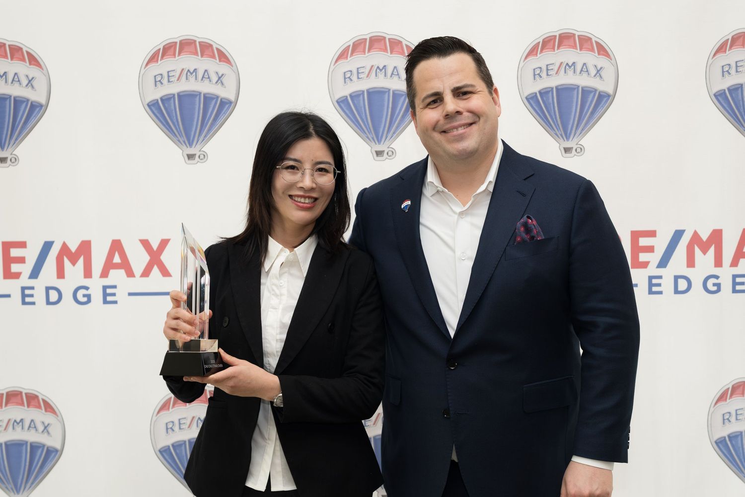 Woman in black blazer proudly holds crystal trophy at RE/MAX Edge real estate awards ceremony.