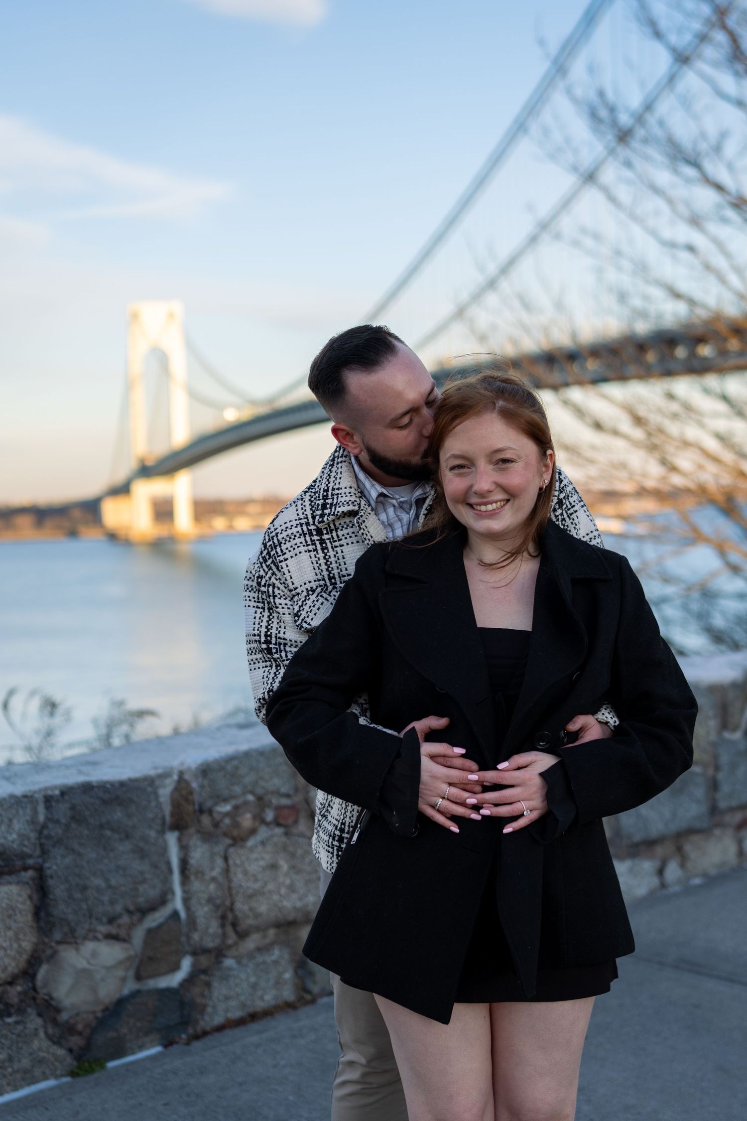 Man kissing woman's cheek from behind as she smiles with the Verrazzano-Narrows Bridge backdrop.