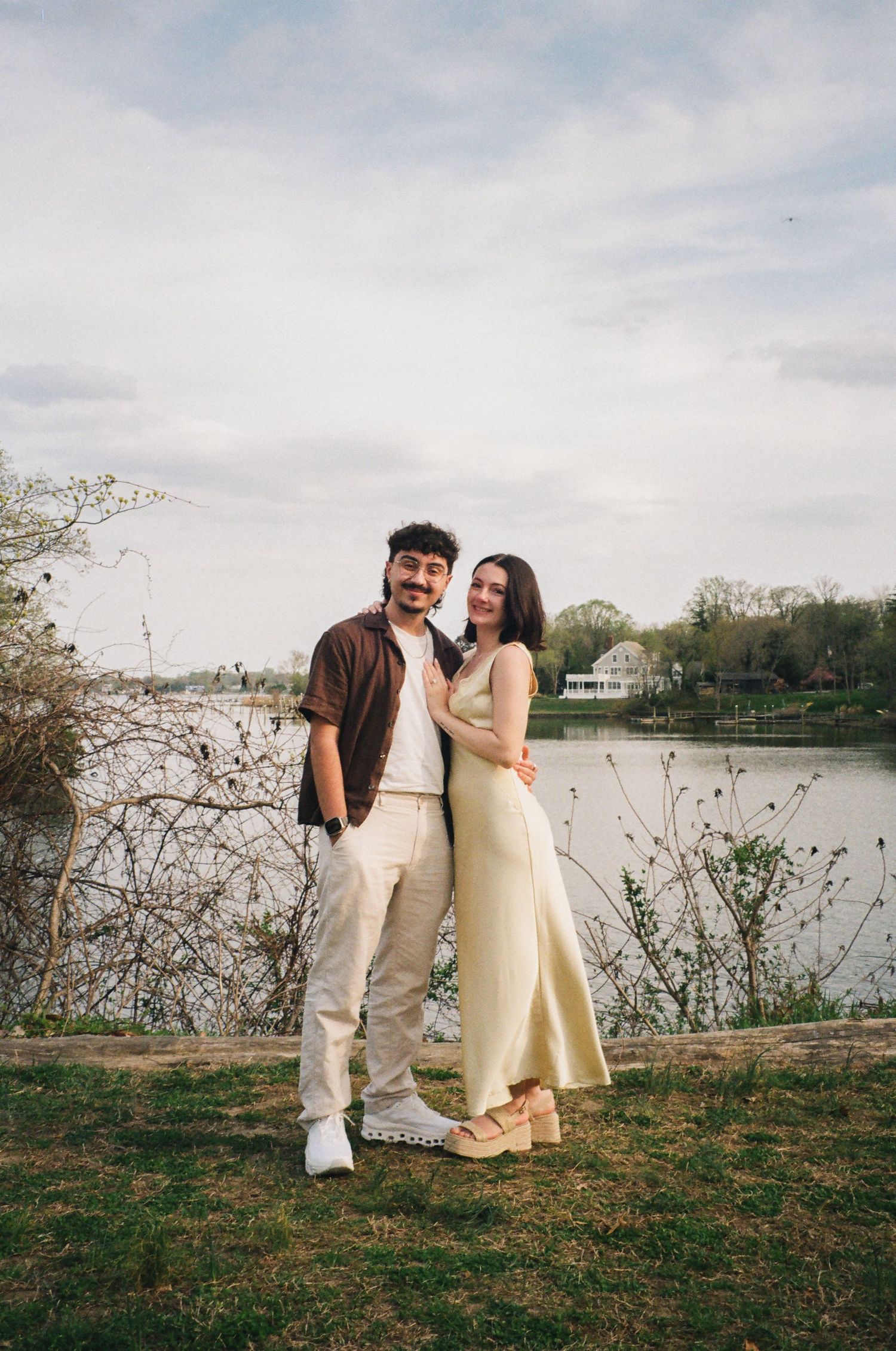 Couple embracing near a serene lake with bare branches and a white house visible across the water.