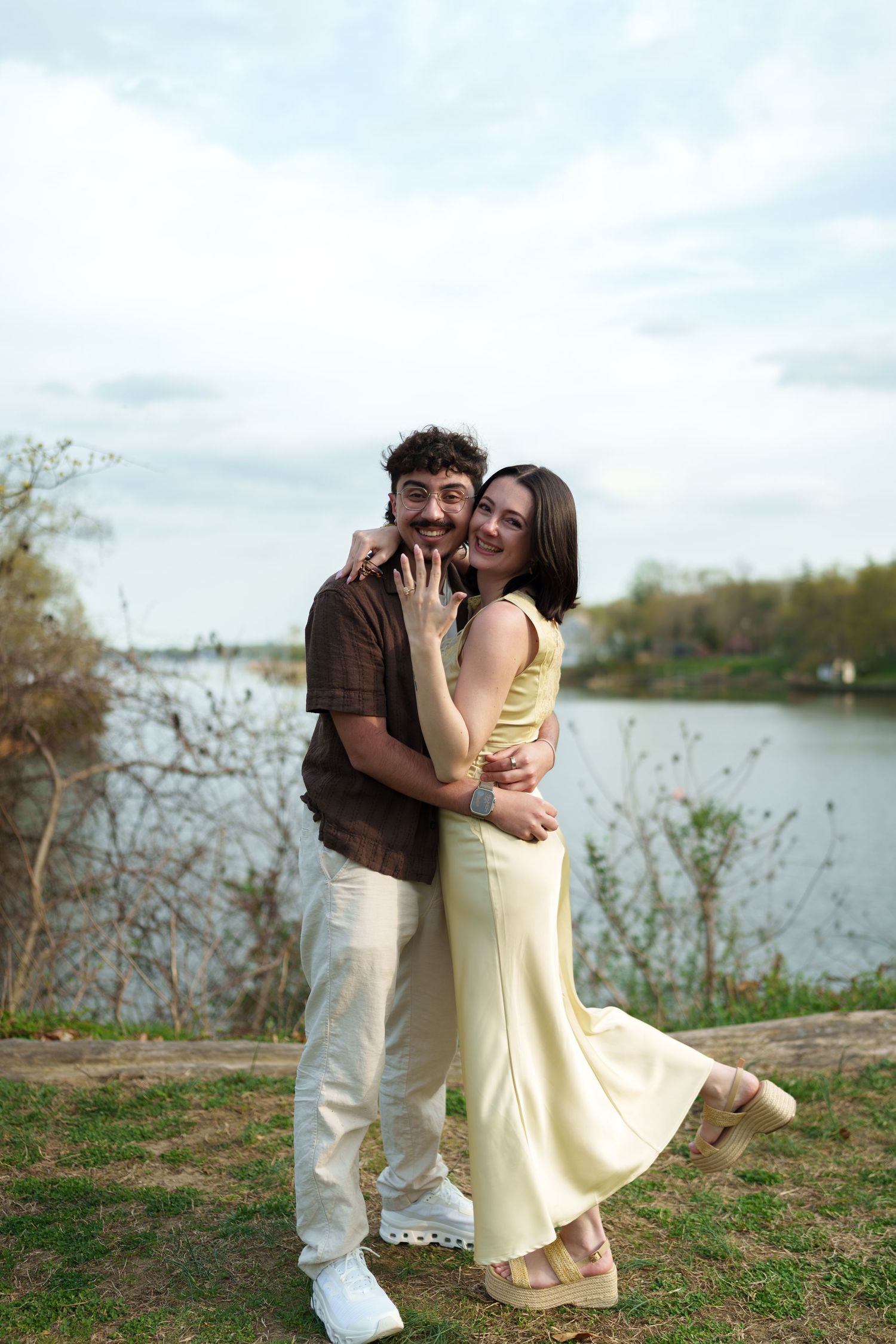 Smiling engaged couple hugging outdoors by a lake, woman showing engagement ring.
