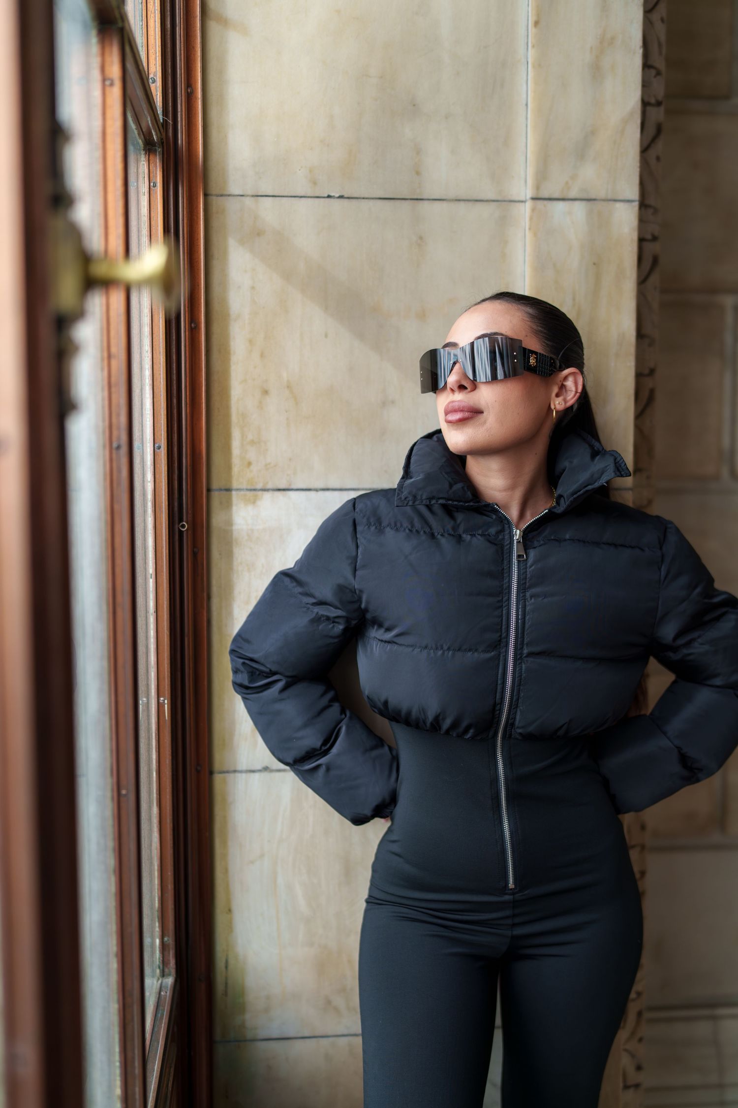 Stylish woman in black puffer jacket and bold sunglasses posing by window near stone wall.