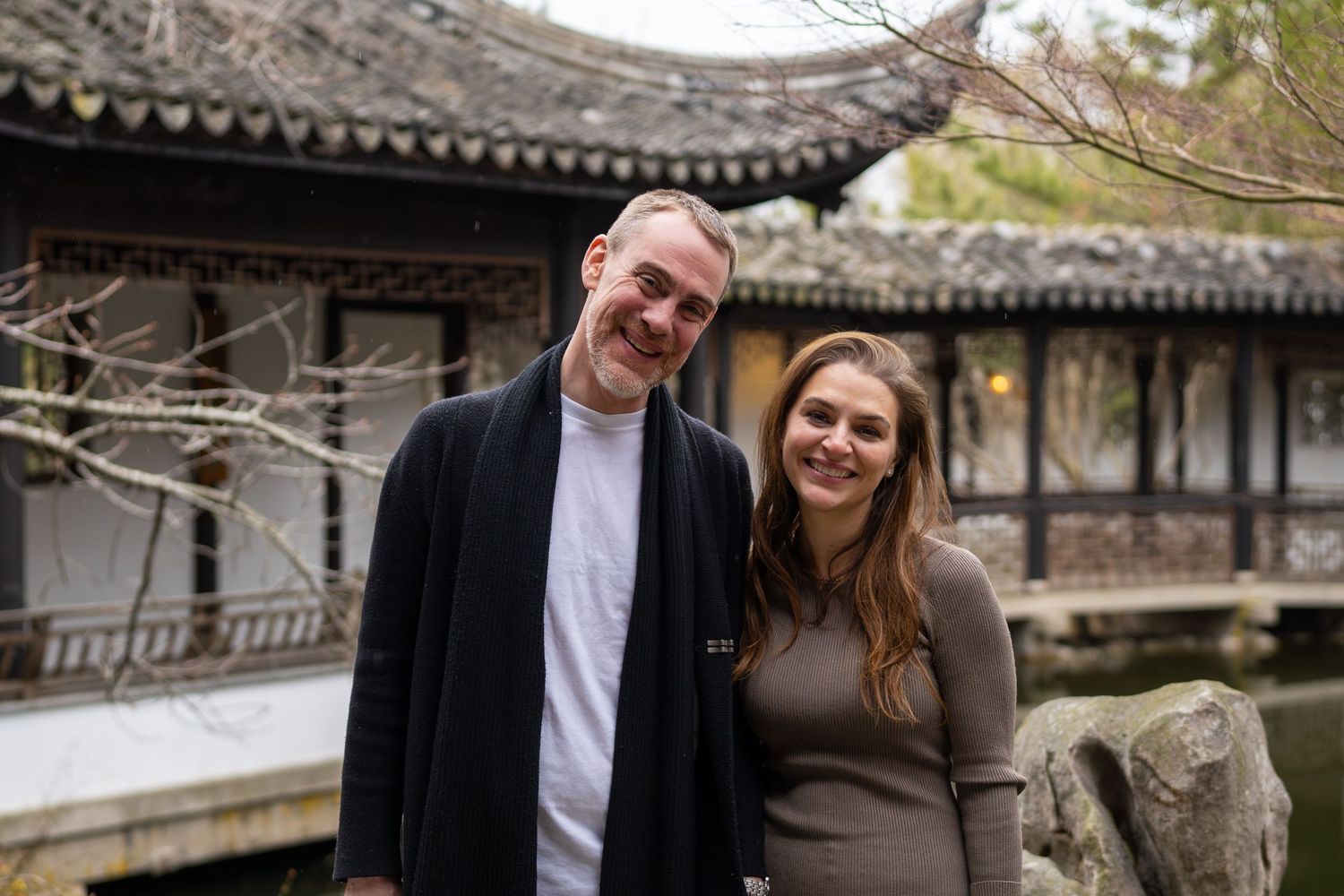 Smiling couple posing together in front of a classic Chinese garden pavilion near a pond.